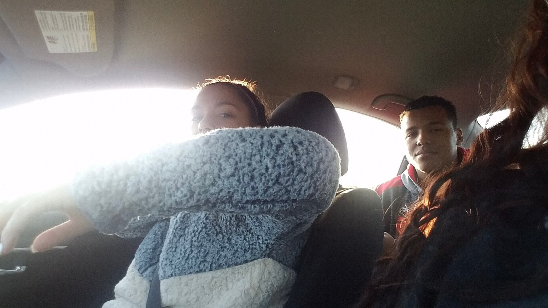 Inside a car with two passengers; a person in the driver’s seat is partially visible and a passenger sits next to them, near a bright window, with a blue-gray fluffy blanket draped over the seat in foreground.