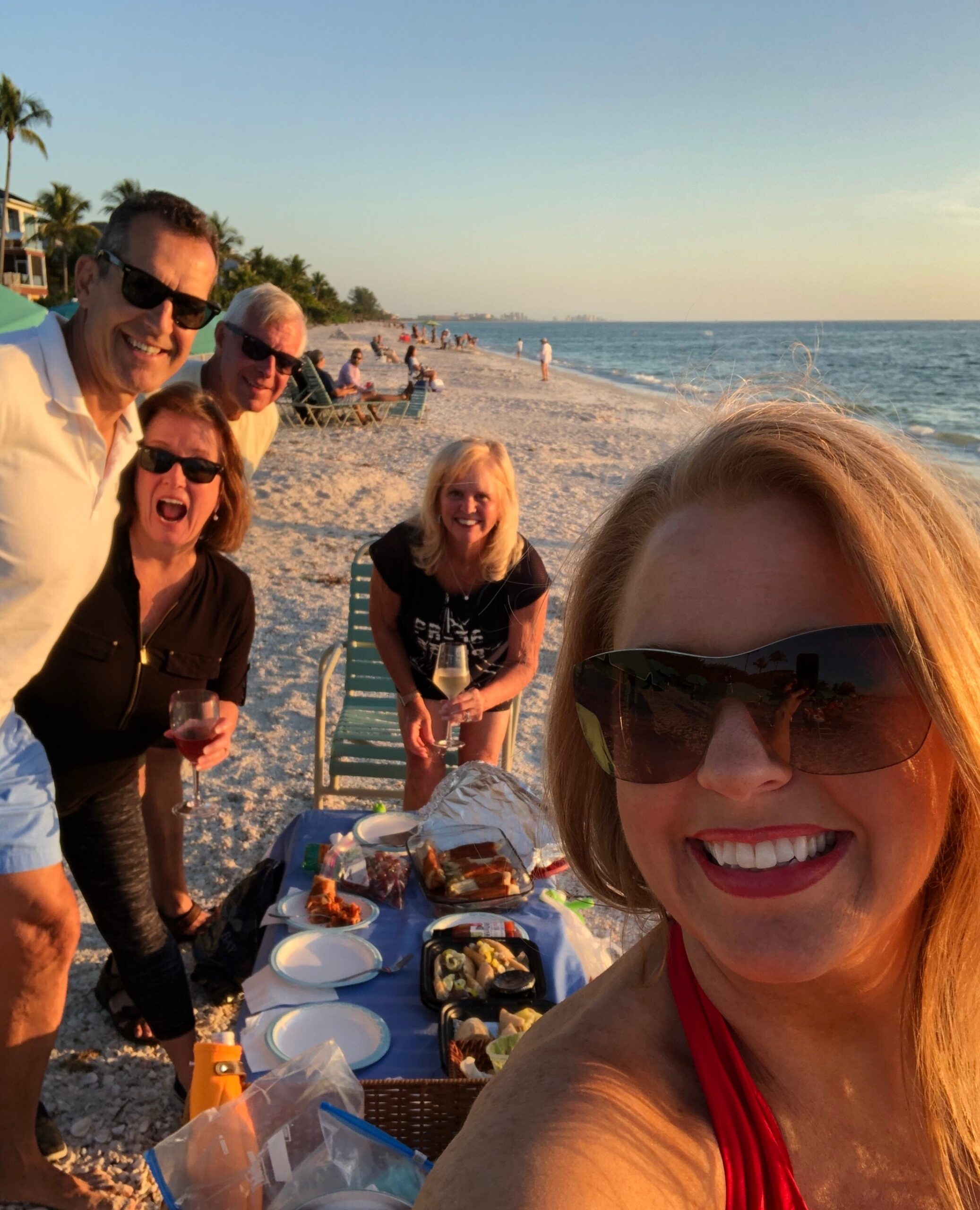 Group of friends on a sunny beach taking a selfie near a table of snacks and drinks, with the ocean in the background.
