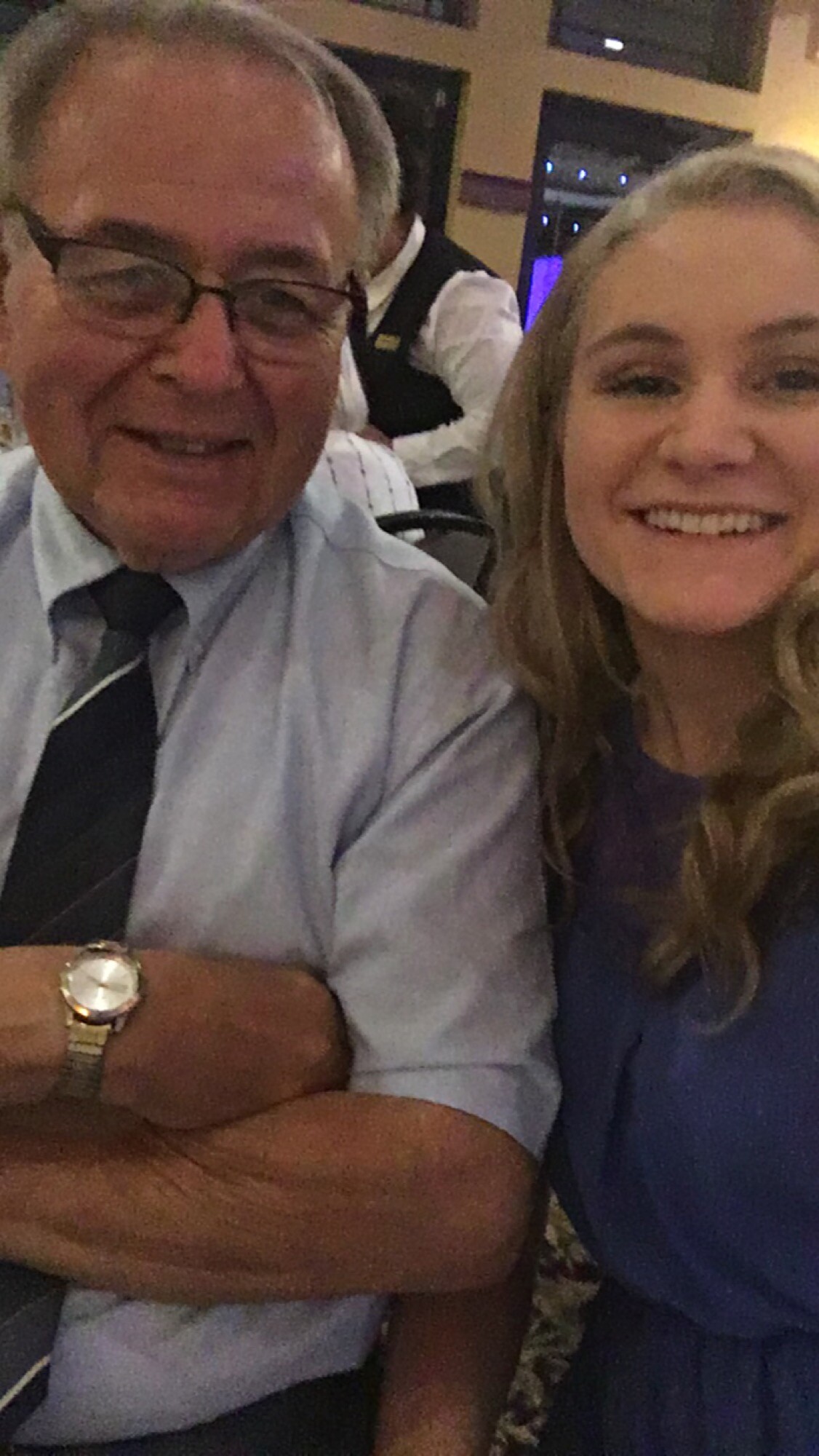Smiling older man in glasses wearing a blue shirt and striped tie sits beside a smiling young woman with long blonde hair at an indoor event.