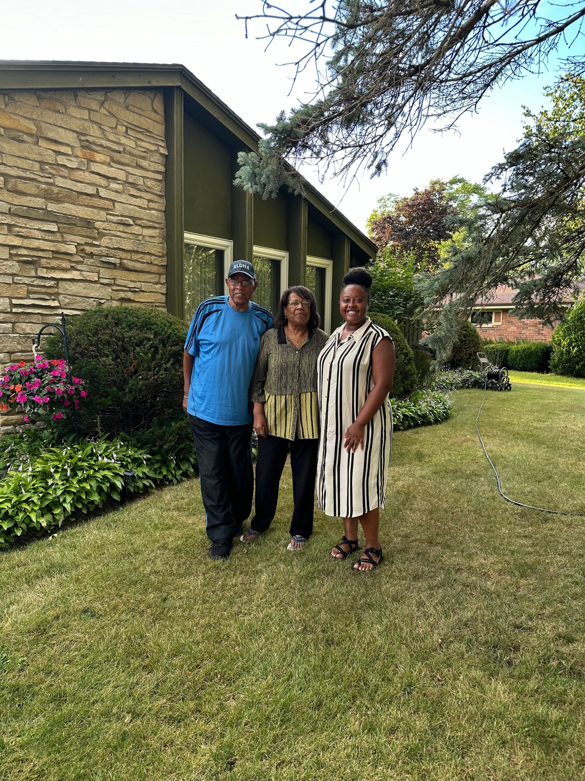 Three people stand together in a front yard in front of a stone and green-painted house, smiling at the camera.