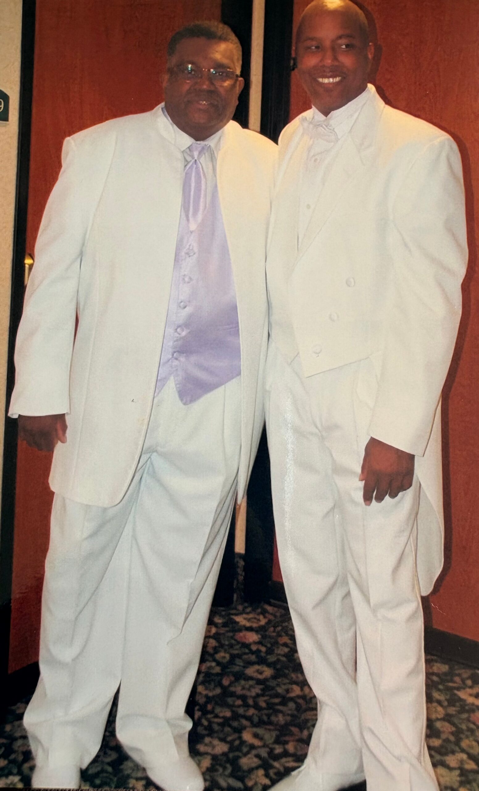 Two men in white tuxedos posing together for a photo at a formal event, standing on a patterned carpet.