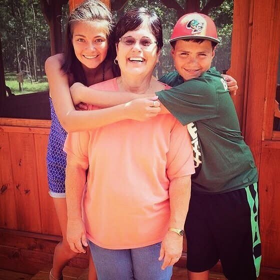 Grandmother smiling as a girl and a boy hug her from both sides outdoors near a wooden structure, on a sunny day.