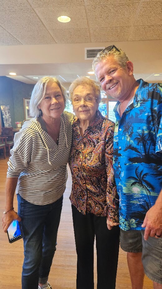 Three adults posing together indoors, smiling for the camera; the woman on the left holds a smartphone in her hand.