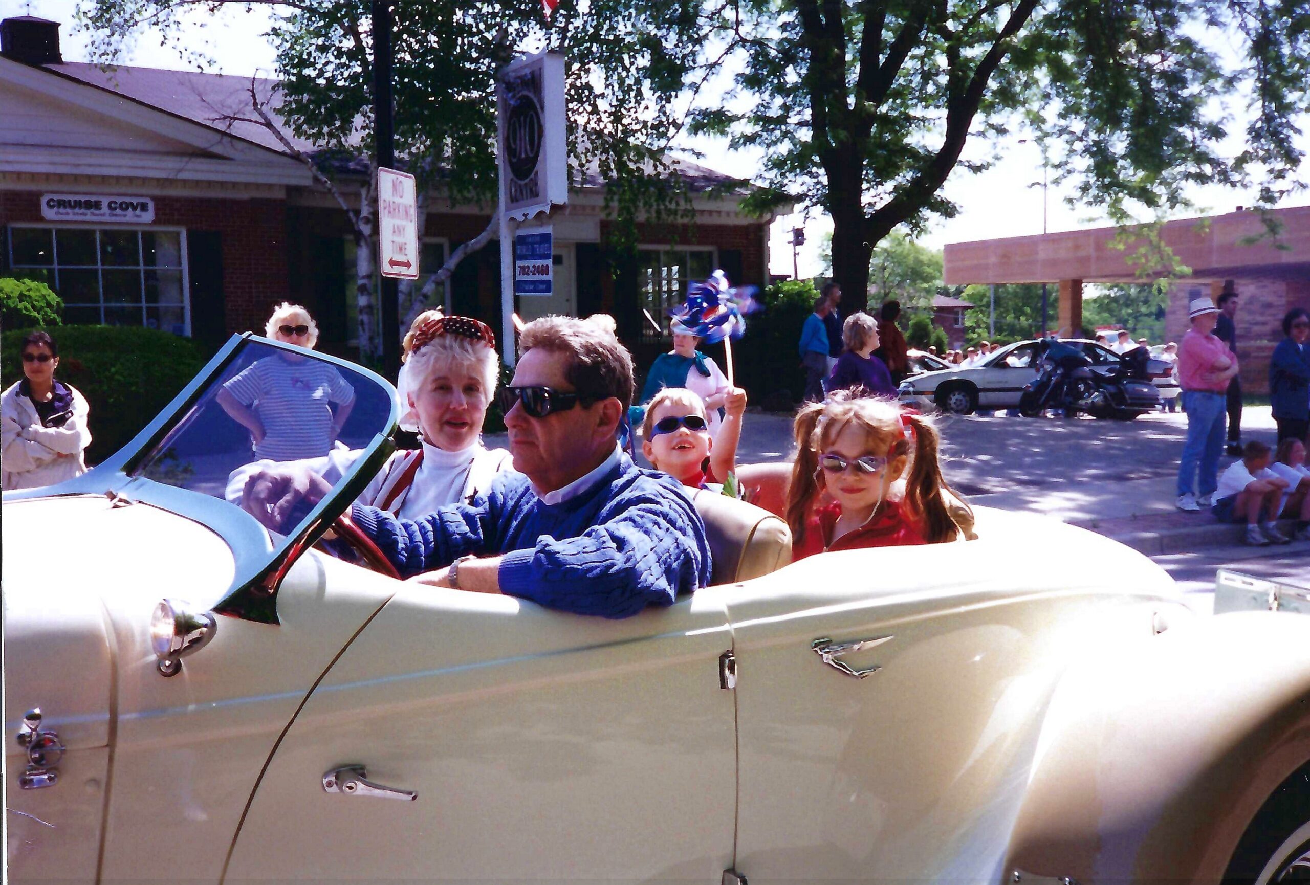 People ride in a light-colored vintage convertible during a daytime parade, with onlookers along the street under a tree.