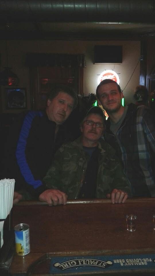 Three men lean on a bar counter in a dimly lit pub, one wearing a camo jacket in front while the other two stand beside him.