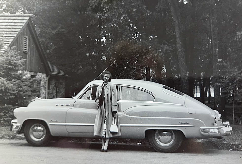 Woman in a long coat poses beside a vintage 1950s car on a driveway in front of a house and trees.