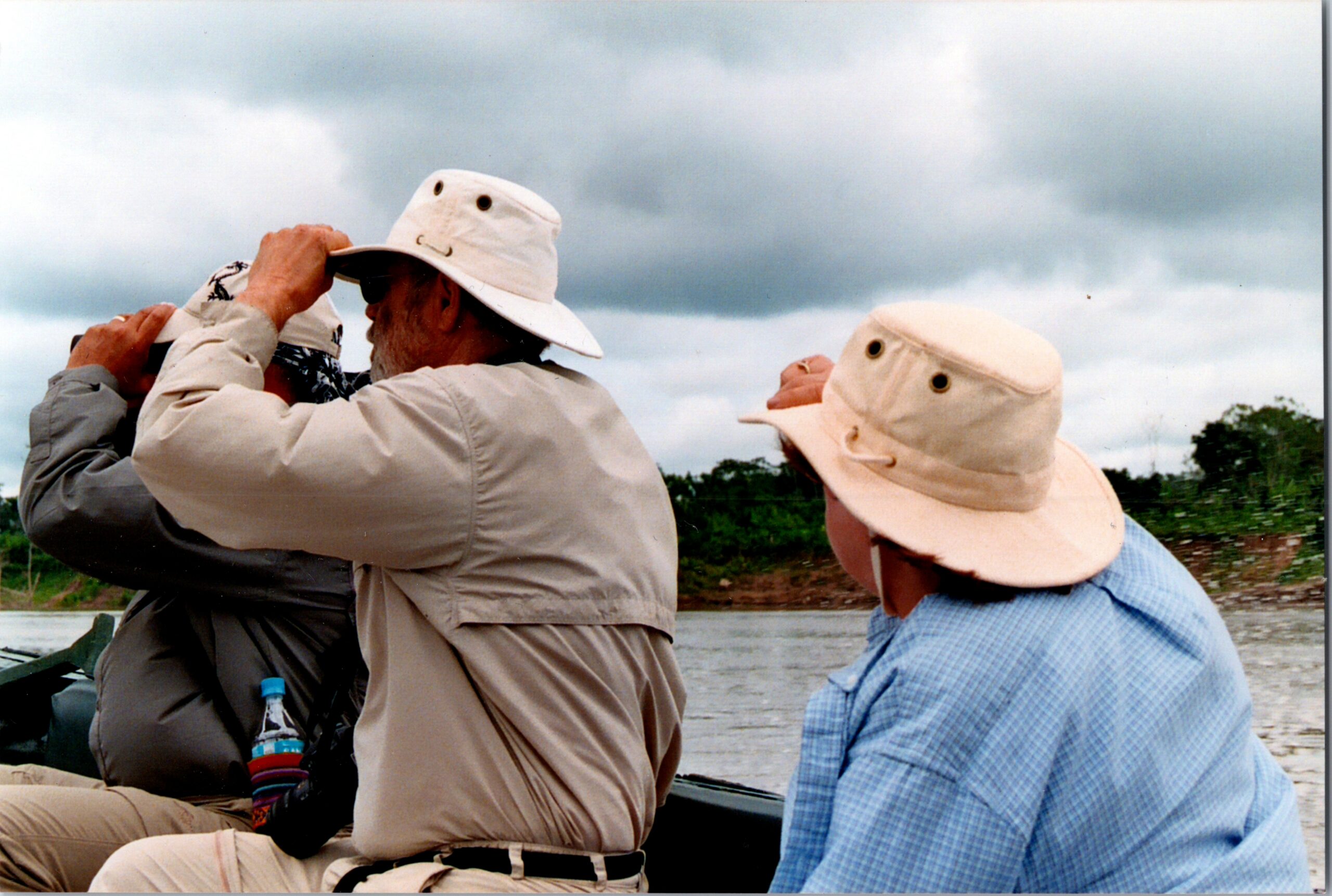 People on a boat wearing wide-brim hats, looking through binoculars toward the water with a cloudy sky above.