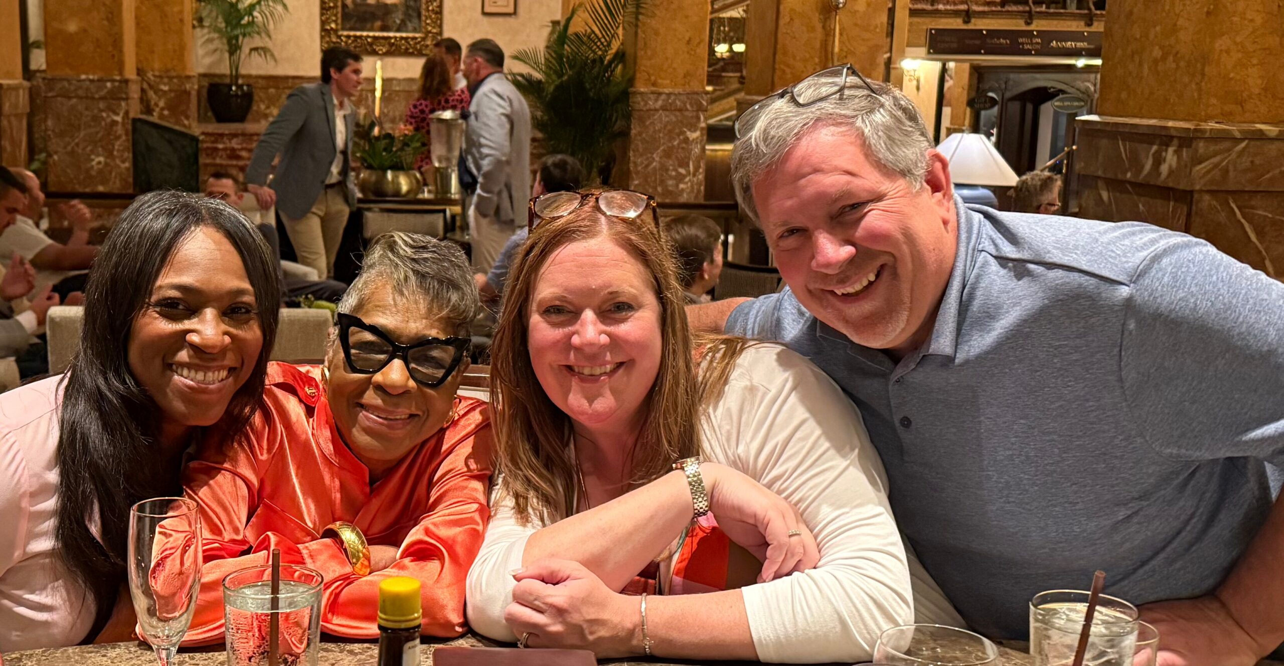 Four friends smiling at a restaurant table, leaning in for a group photo with drinks in front.