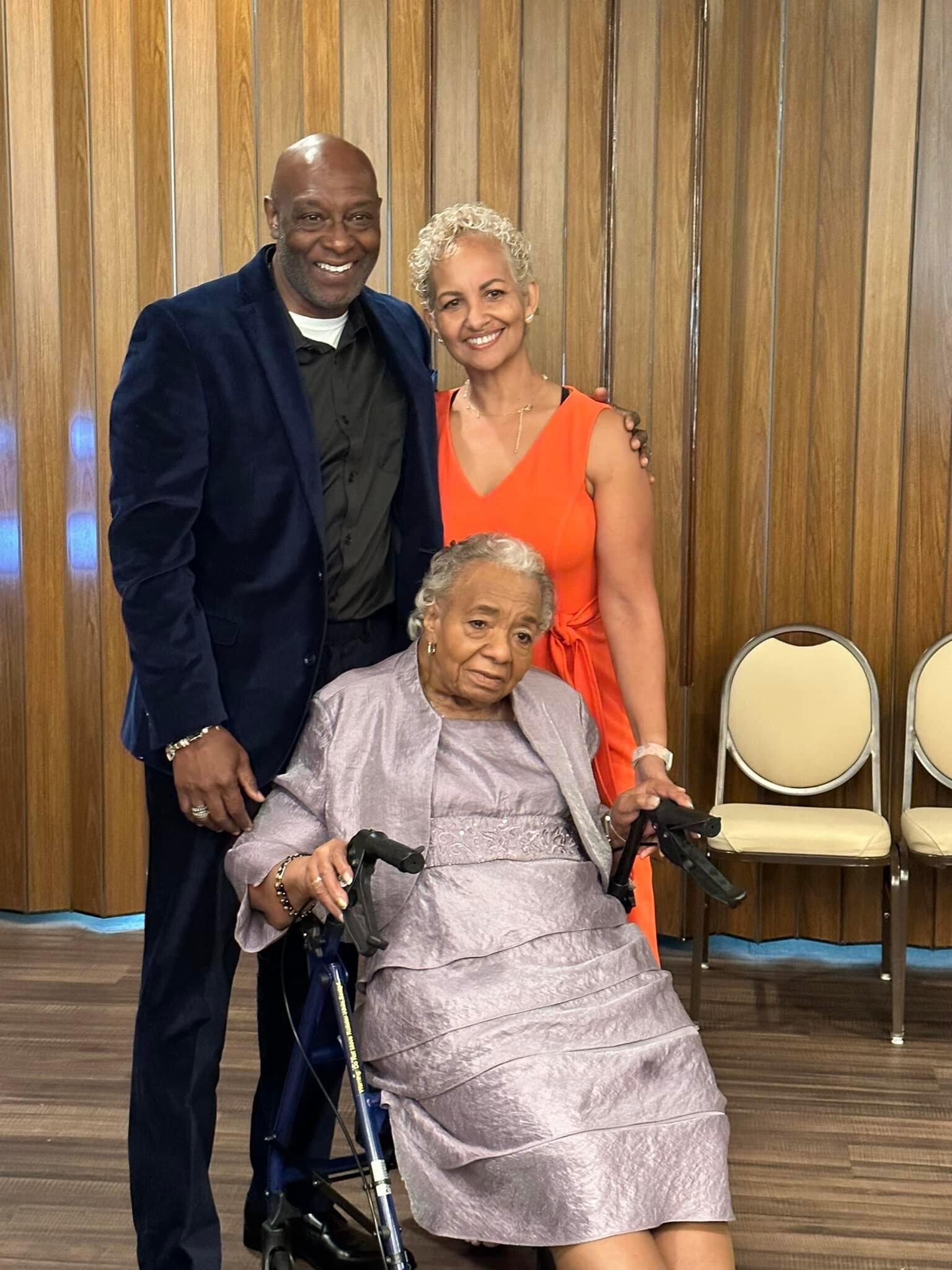 Three people posing indoors: a man in a dark navy suit, a woman in an orange dress, and an elderly woman in a wheelchair in front of them.