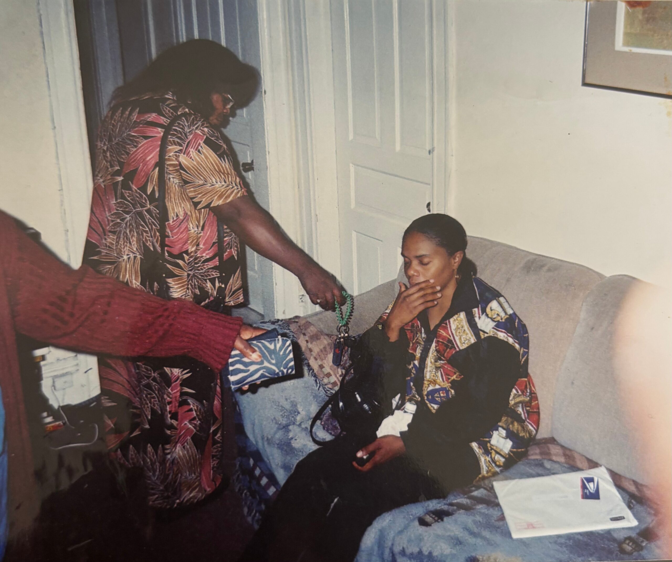 Older person in a bright tropical shirt hands a green bead necklace to a seated man on a sofa, in a living room setting.