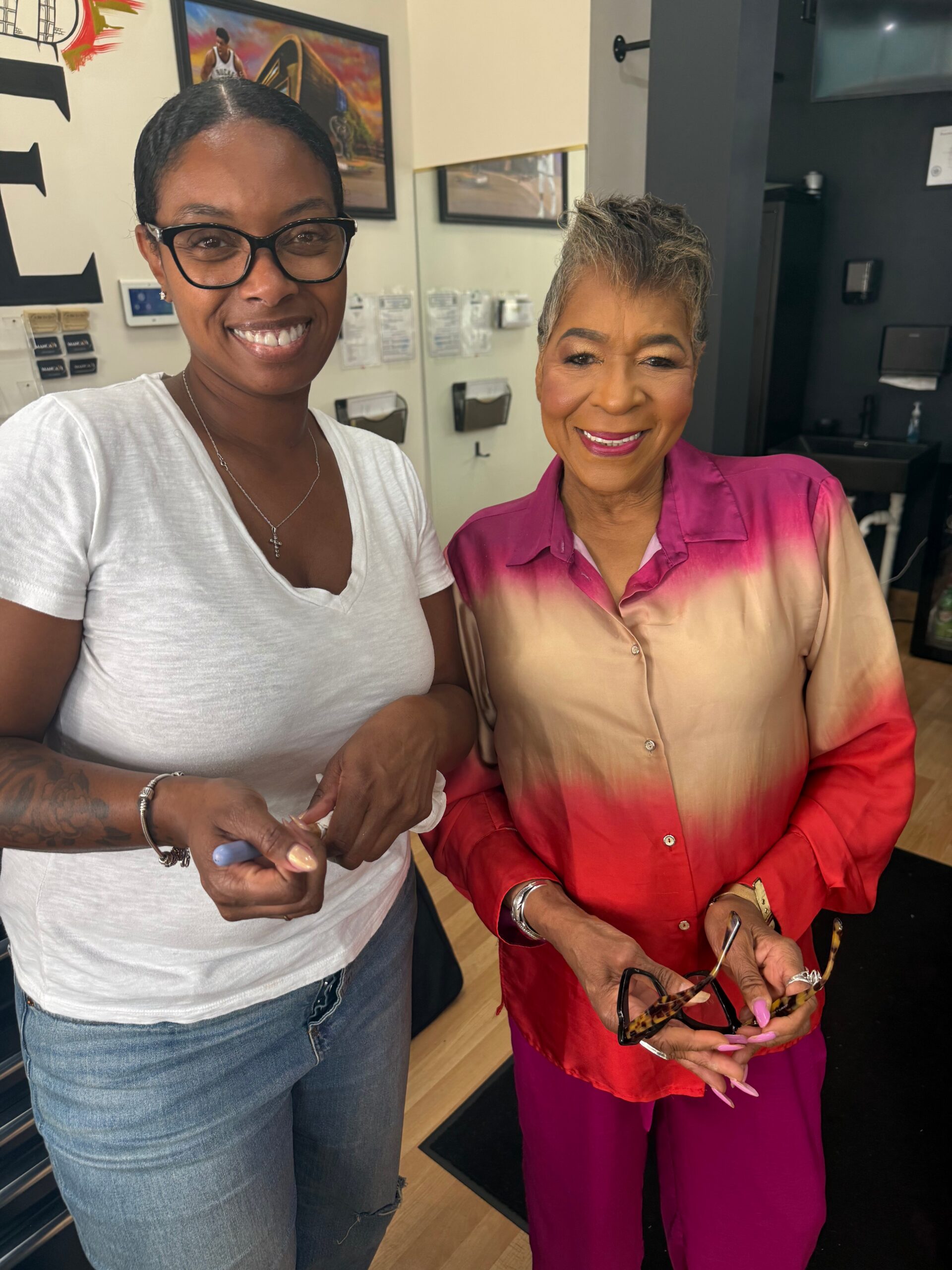 Two women stand side by side in a nail salon, smiling; left wears a white shirt and glasses, right wears a pink gradient shirt and holds nail tools.