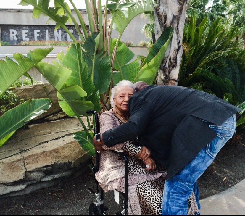 Elderly woman in a wheelchair receiving a hug from a man outdoors among tropical plants.
