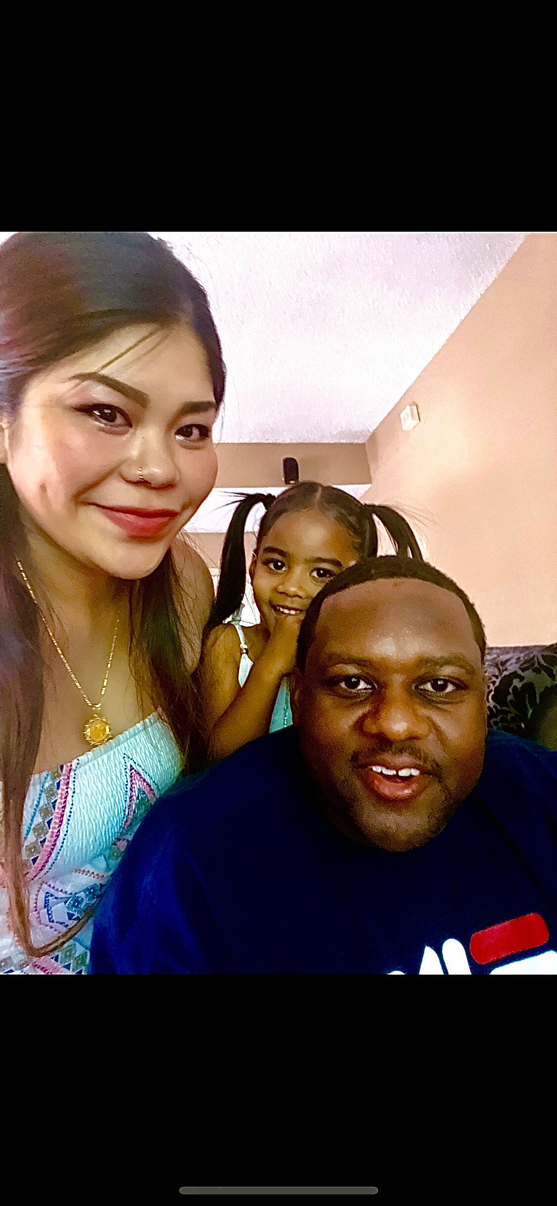 Three people pose for a selfie indoors: a smiling woman on the left, a man in the foreground on the right, and a girl between them smiling at the camera.