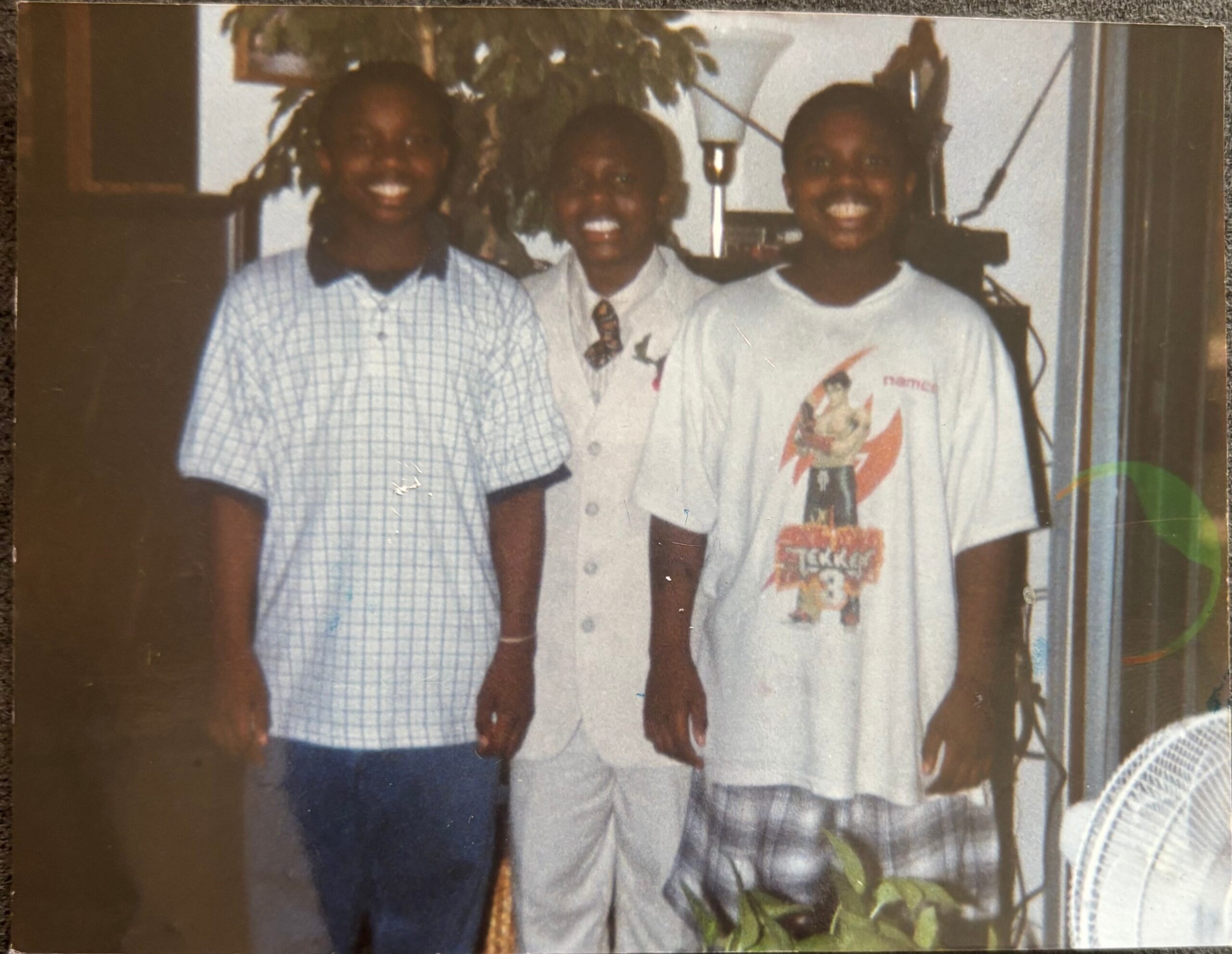 Three smiling friends posing together indoors, standing side by side in front of a plant and lamp behind them.