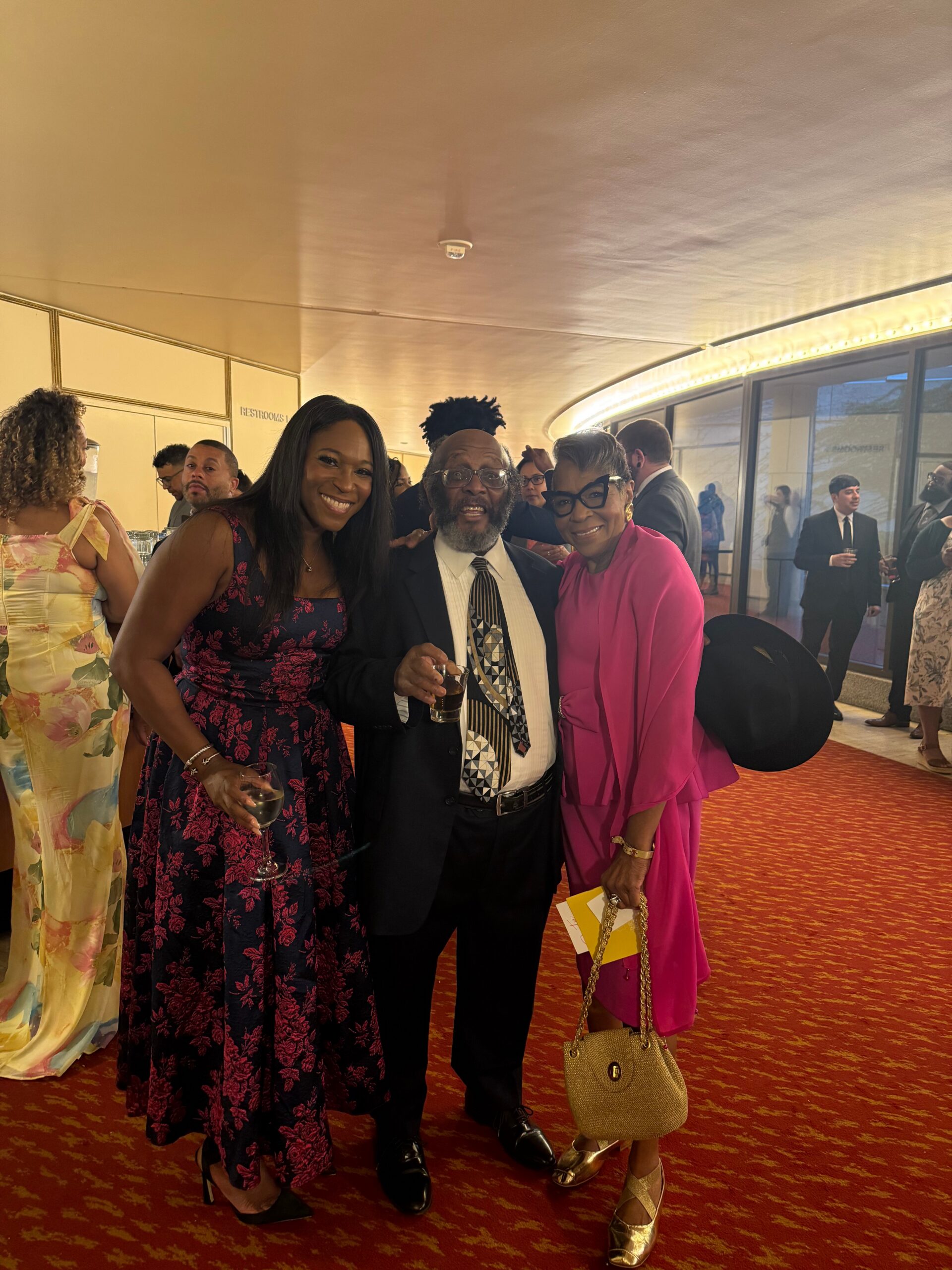 Three smiling guests posing together at a formal event, two women flanking a man in a suit, each holding drinks in a banquet hall.