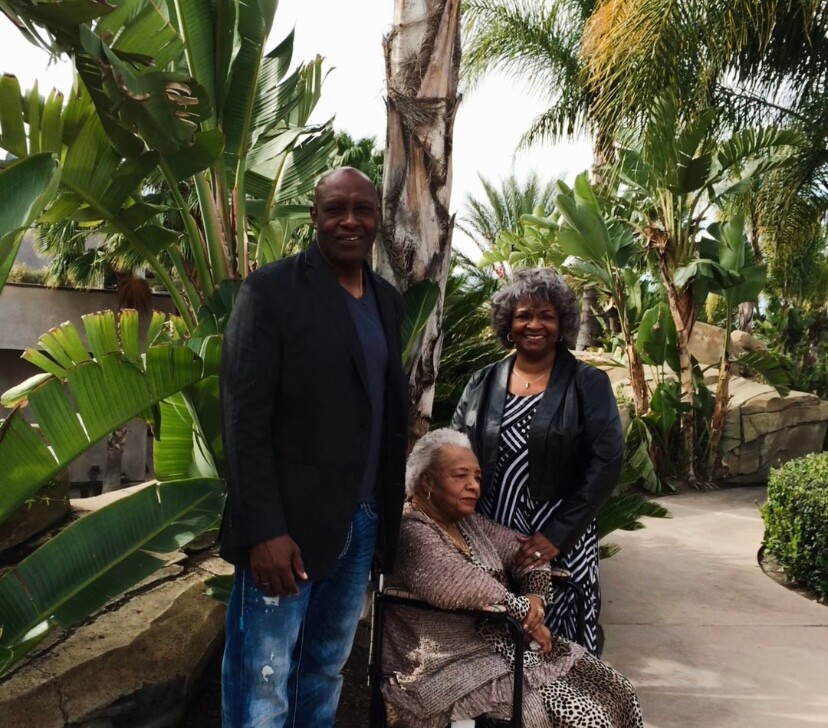 Three generations posing outdoors: an elderly woman in a wheelchair flanked by a man and a woman among tropical plants.