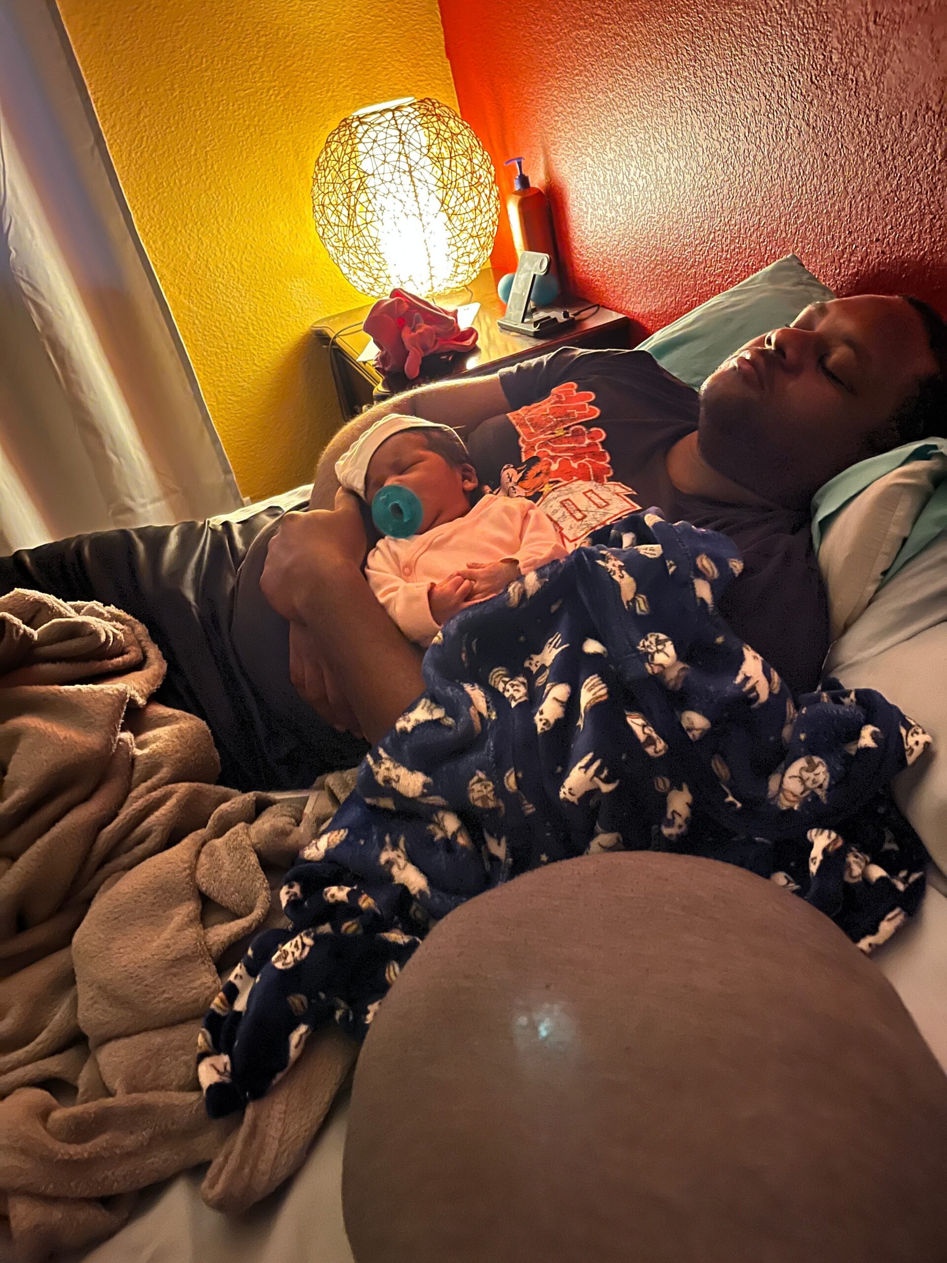 Man lying in bed cradling a newborn baby with a blue pacifier, wrapped in a patterned navy blanket under warm lighting.