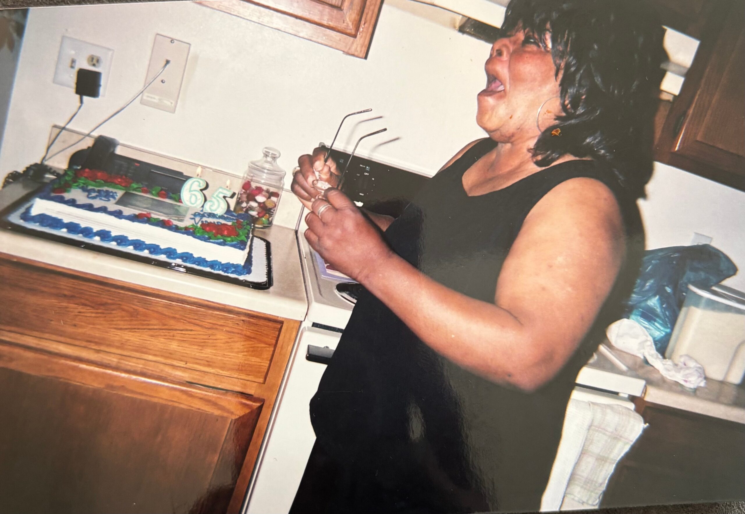 Woman in a black sleeveless top blowing out candles on a rectangular birthday cake with blue icing, the cake showing a 65 candle in a kitchen setting.
