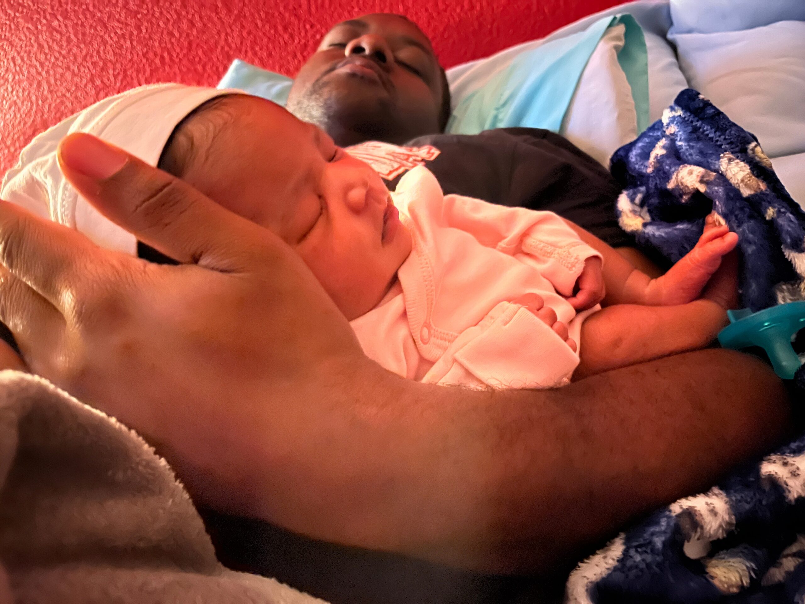 Newborn baby sleeping in a caregiver's arms with a pink outfit and a blue blanket nearby against a red wall.