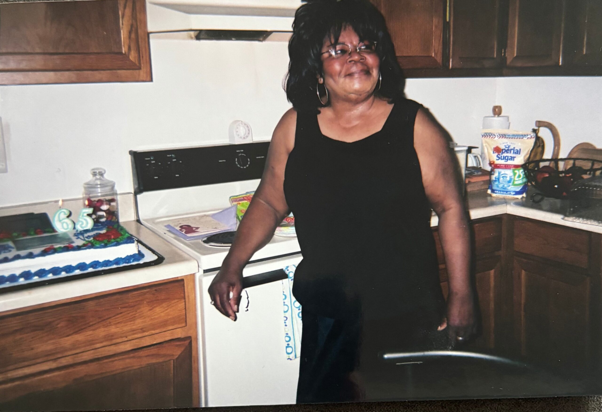 Smiling woman in a black dress standing in a kitchen beside a birthday cake with lit '65' candles