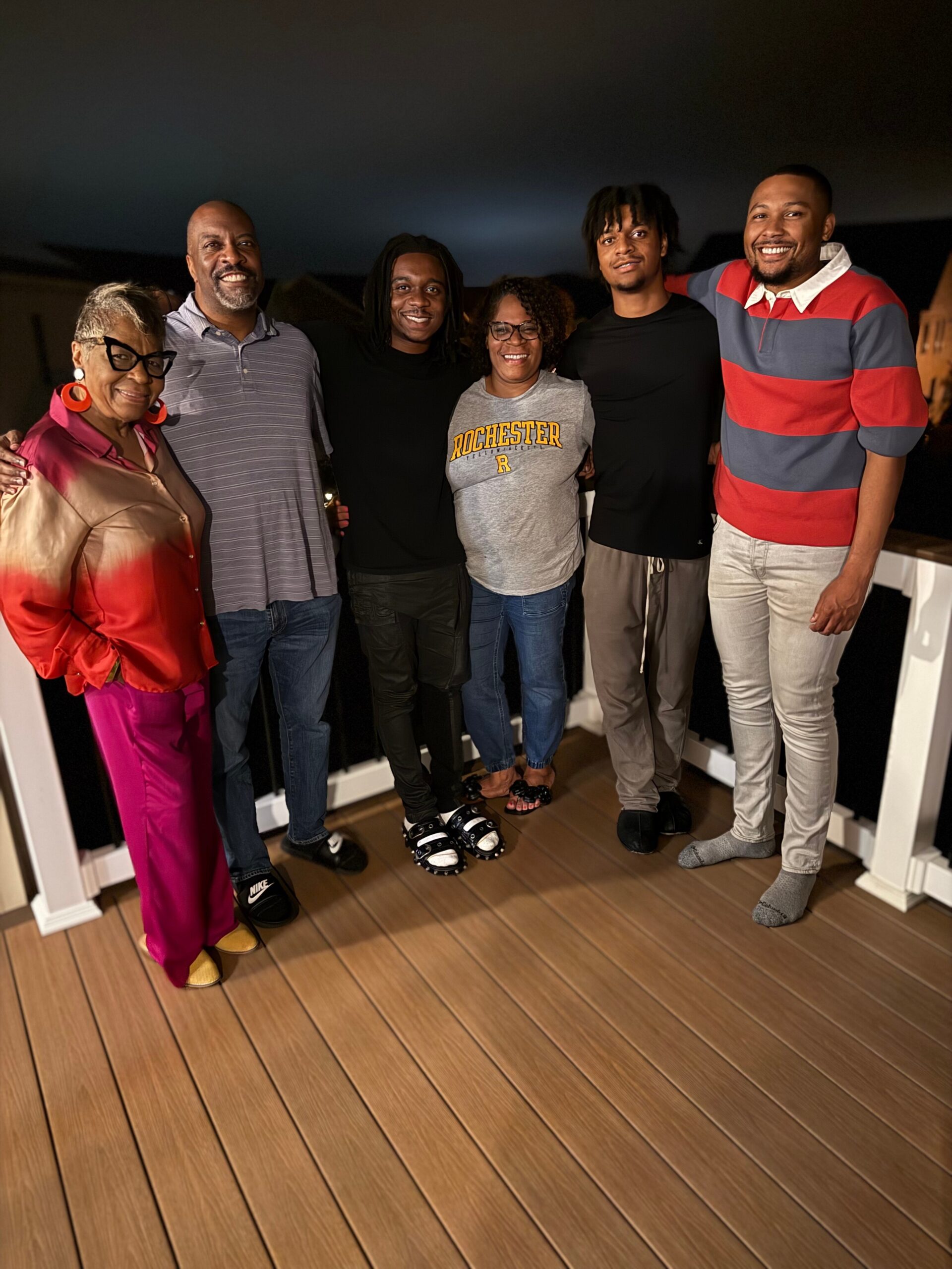 Six adults of diverse ages stand shoulder to shoulder on a wooden balcony at night, smiling at the camera.