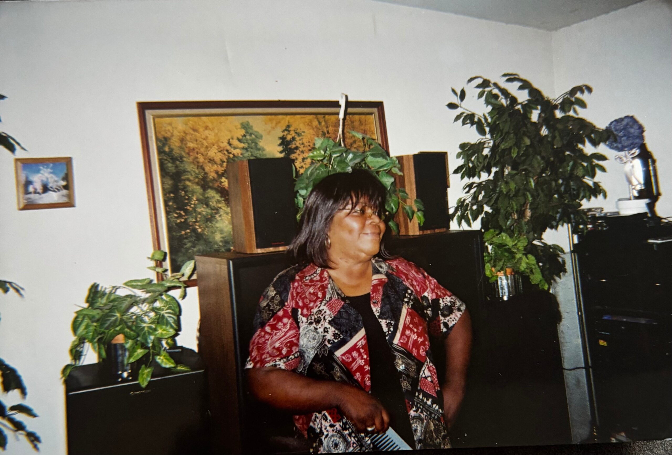 Smiling woman with dark hair in a red patterned blouse sits at a desk in a plant-filled living room. A framed picture and large green plants are behind her.