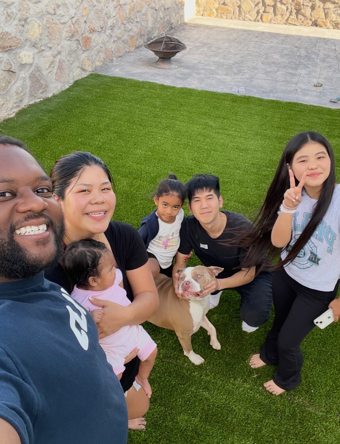 Group of six people and a brown-and-white dog posing for a selfie on a green lawn beside a stone wall in a backyard.