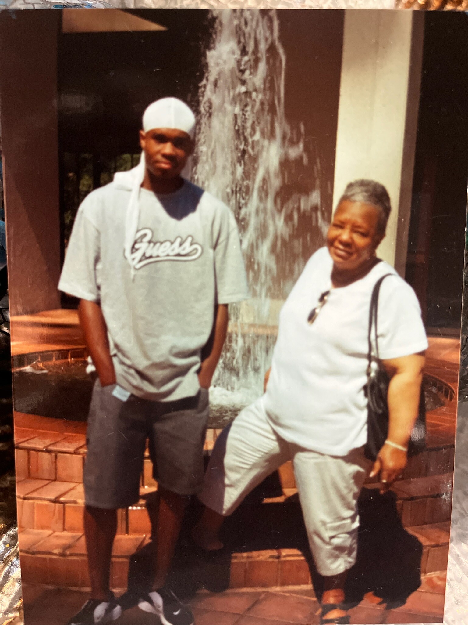 Two people standing near a fountain outdoors; man in a gray shirt with a 'Guess' logo and a white headwrap, woman in white with a black bag.
