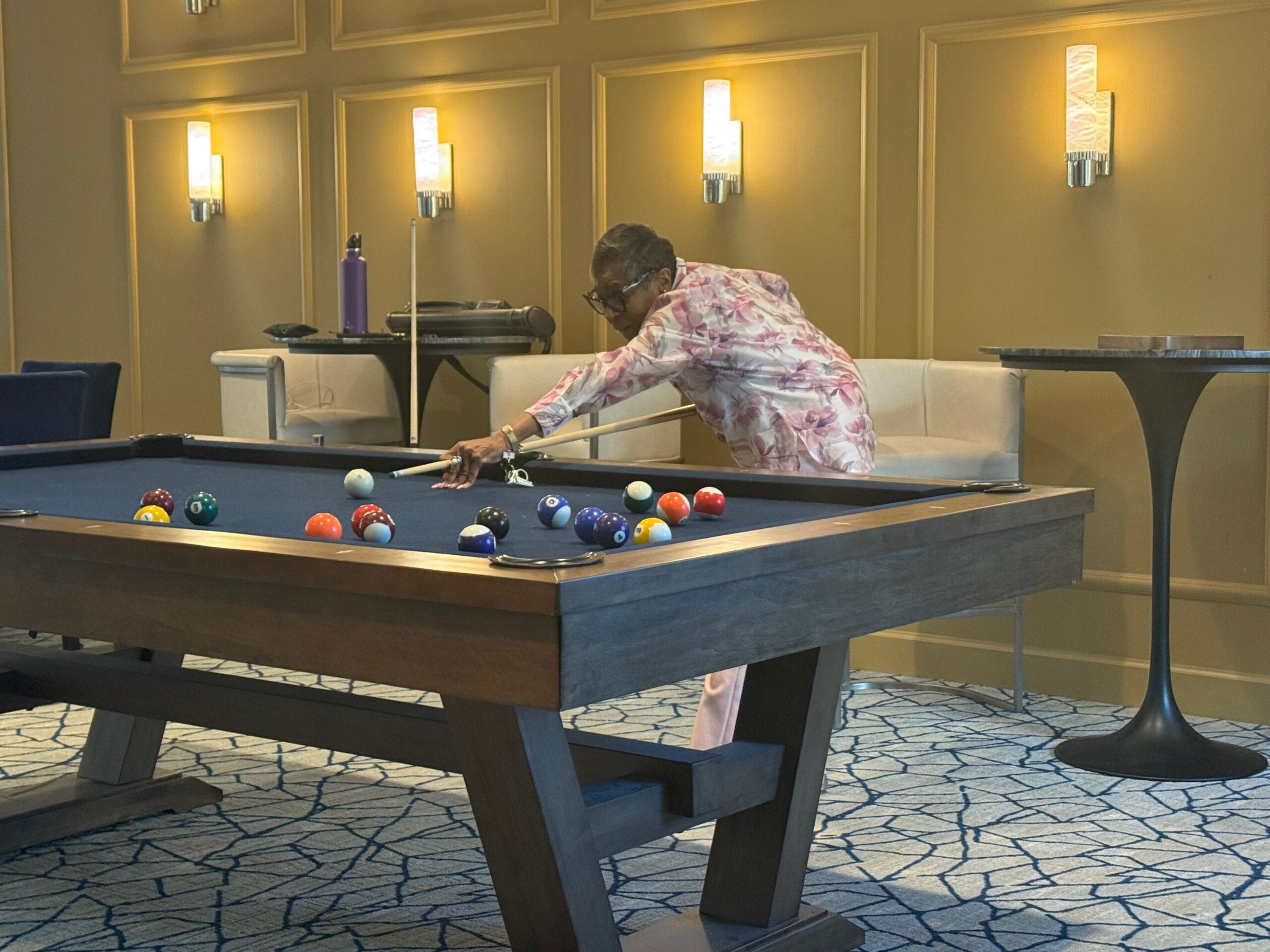 Older woman in a pink floral shirt leans over a pool table to take a shot in a lounge setting.
