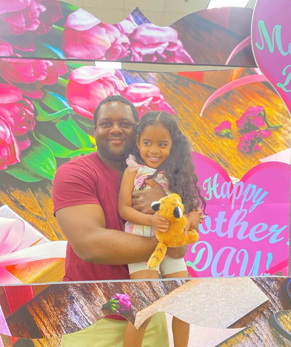 Man holding a smiling girl in a colorful Mother’s Day backdrop with pink flowers; girl holds a yellow plush toy.