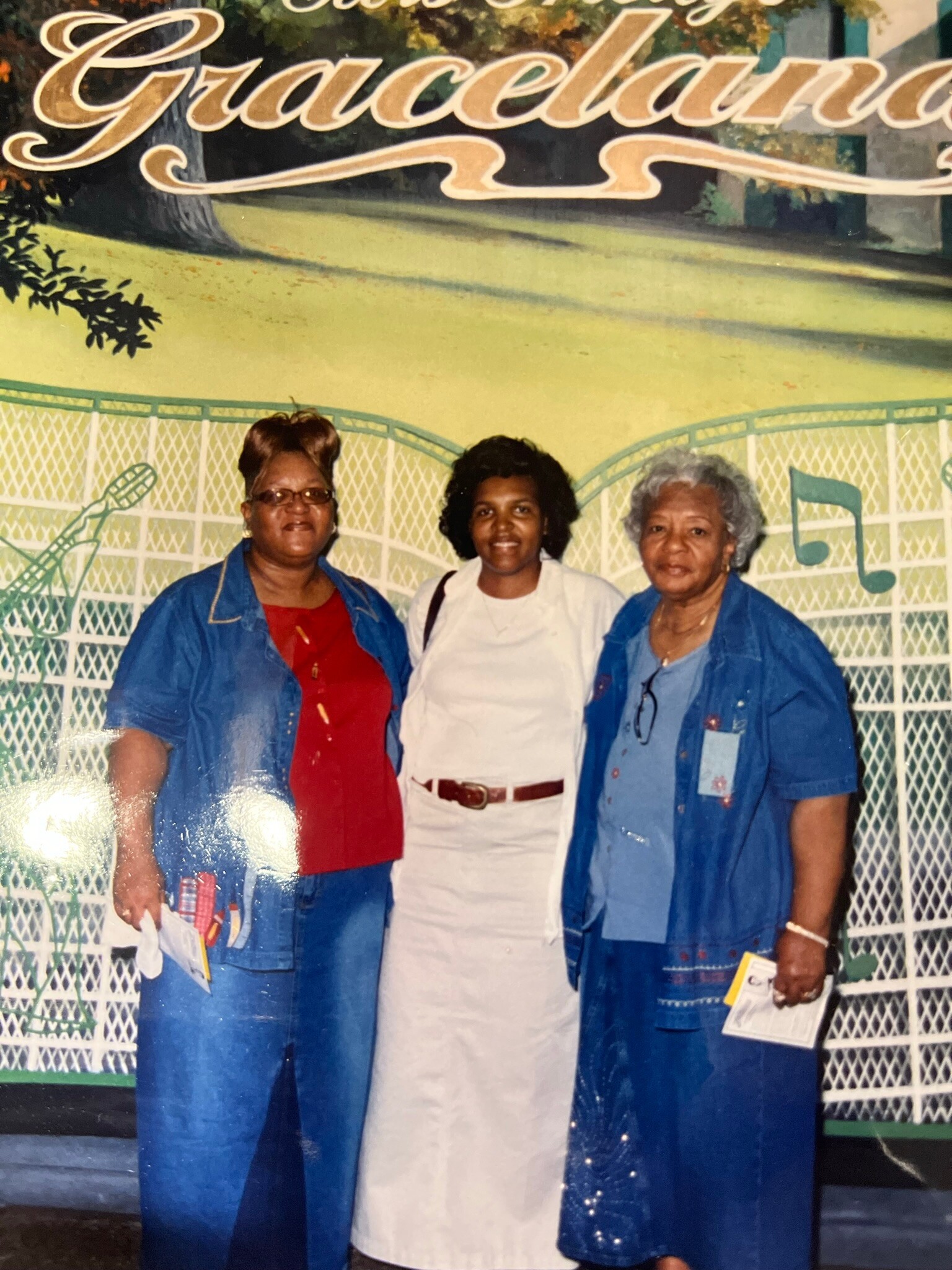 Three women pose in front of a Graceland mural; two wear blue jackets and one wears a white dress.