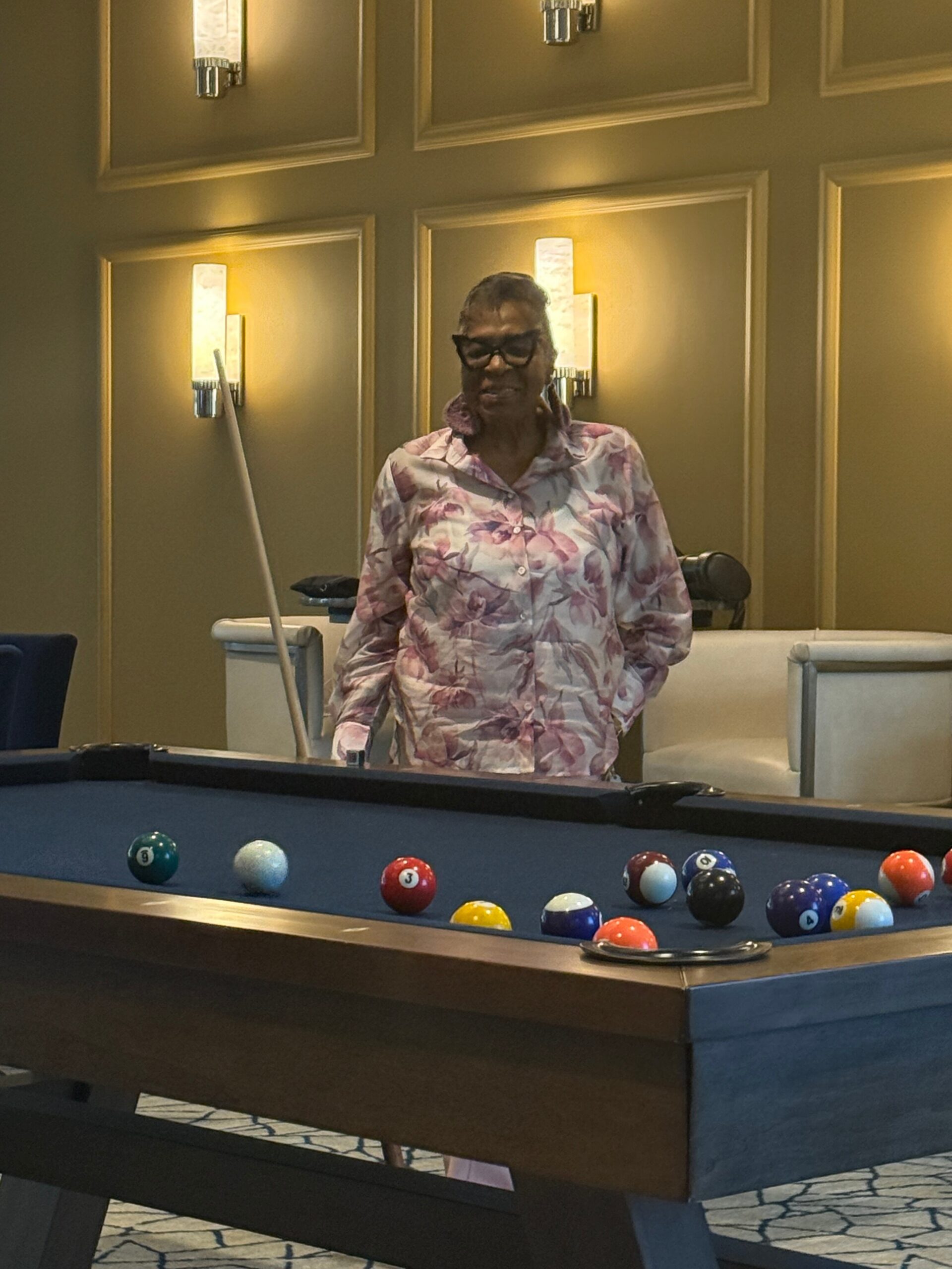 Person wearing a pink floral shirt and glasses stands at a pool table with billiard balls on the felt in a warmly lit room.