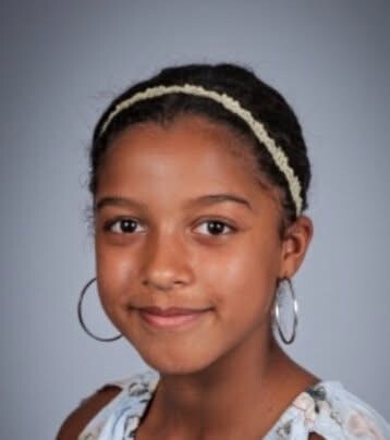 Portrait of a young girl with dark hair, wearing a cream headband and large hoop earrings, smiling softly at the camera.