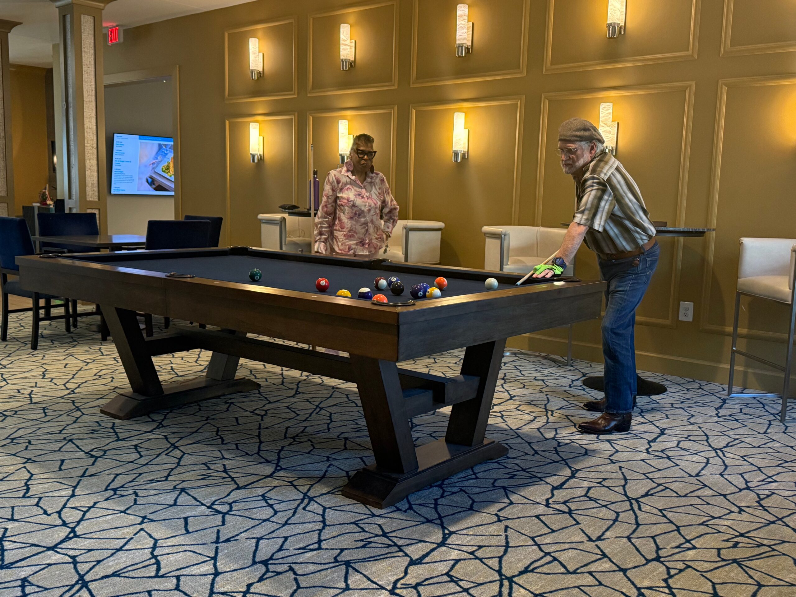 Two people playing pool in a luxe lounge; man in a plaid shirt and gray cap lines up a shot as a woman in a floral blouse watches.