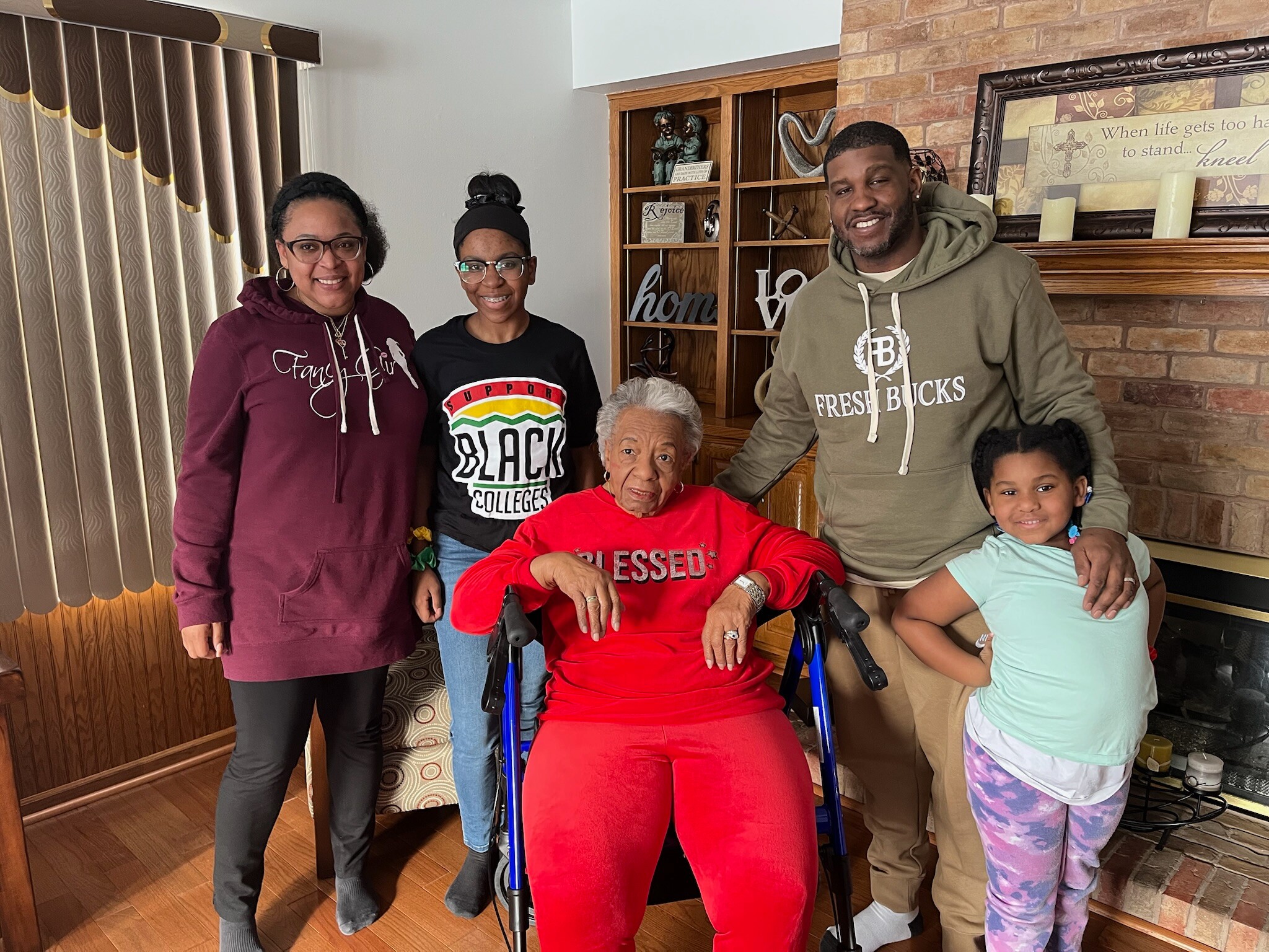 Six people pose together in a living room, including two women on the left, a woman in red seated in a wheelchair center, a man in a hoodie on the right, and a young girl on the far right beside him.