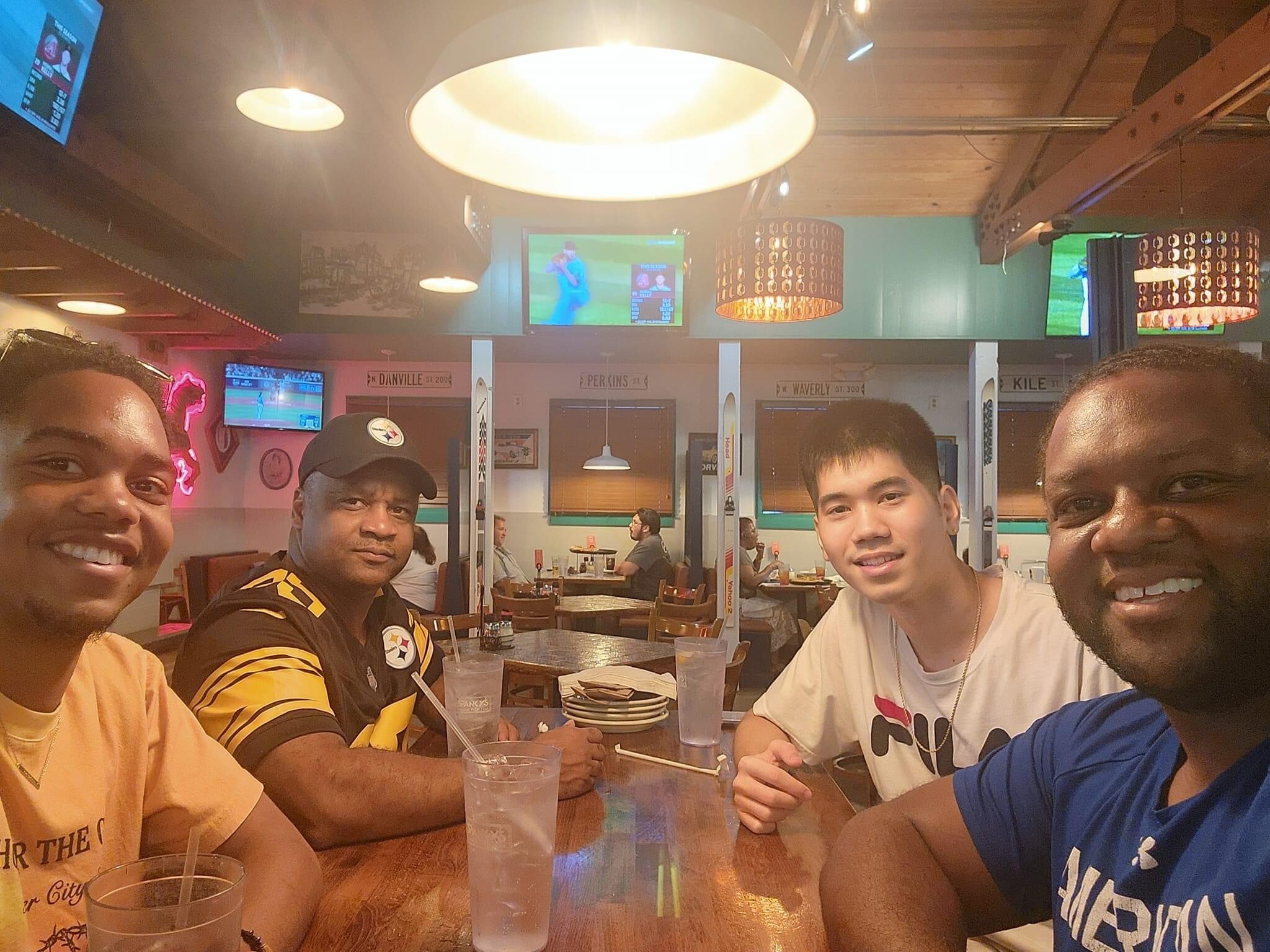 Four friends sit at a wooden table in a casual sports bar, drinks in plastic cups, smiling at the camera.