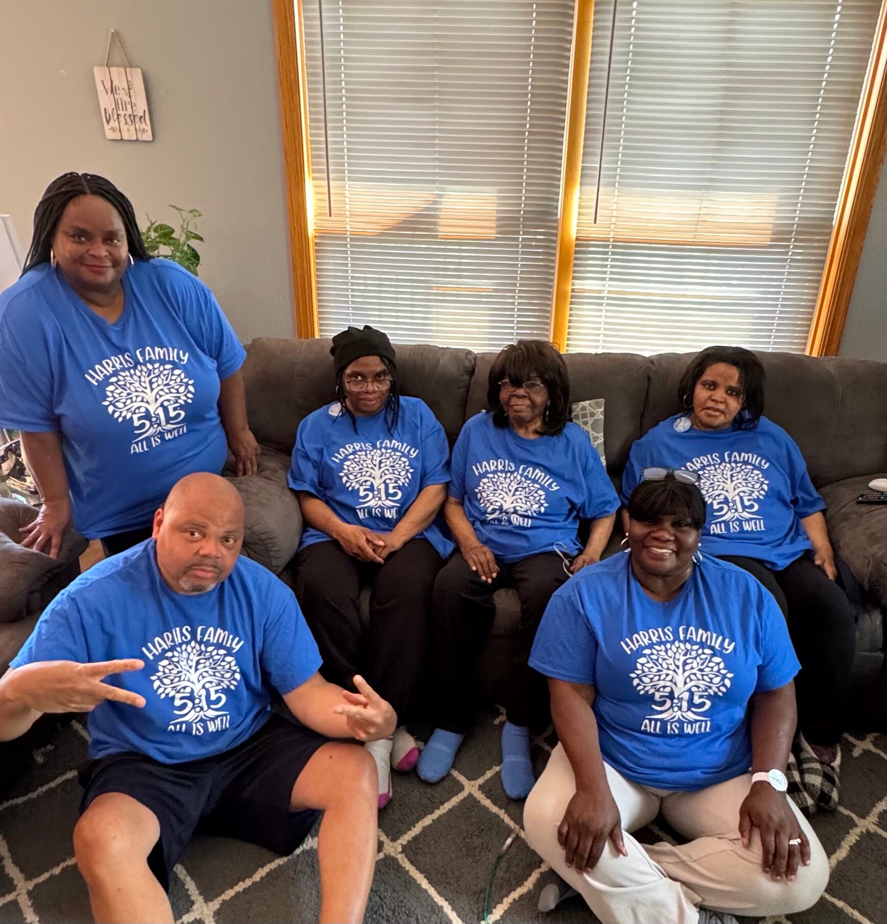 Seven family members in matching blue Harris Family shirts posing on a living room sofa with window blinds behind them.