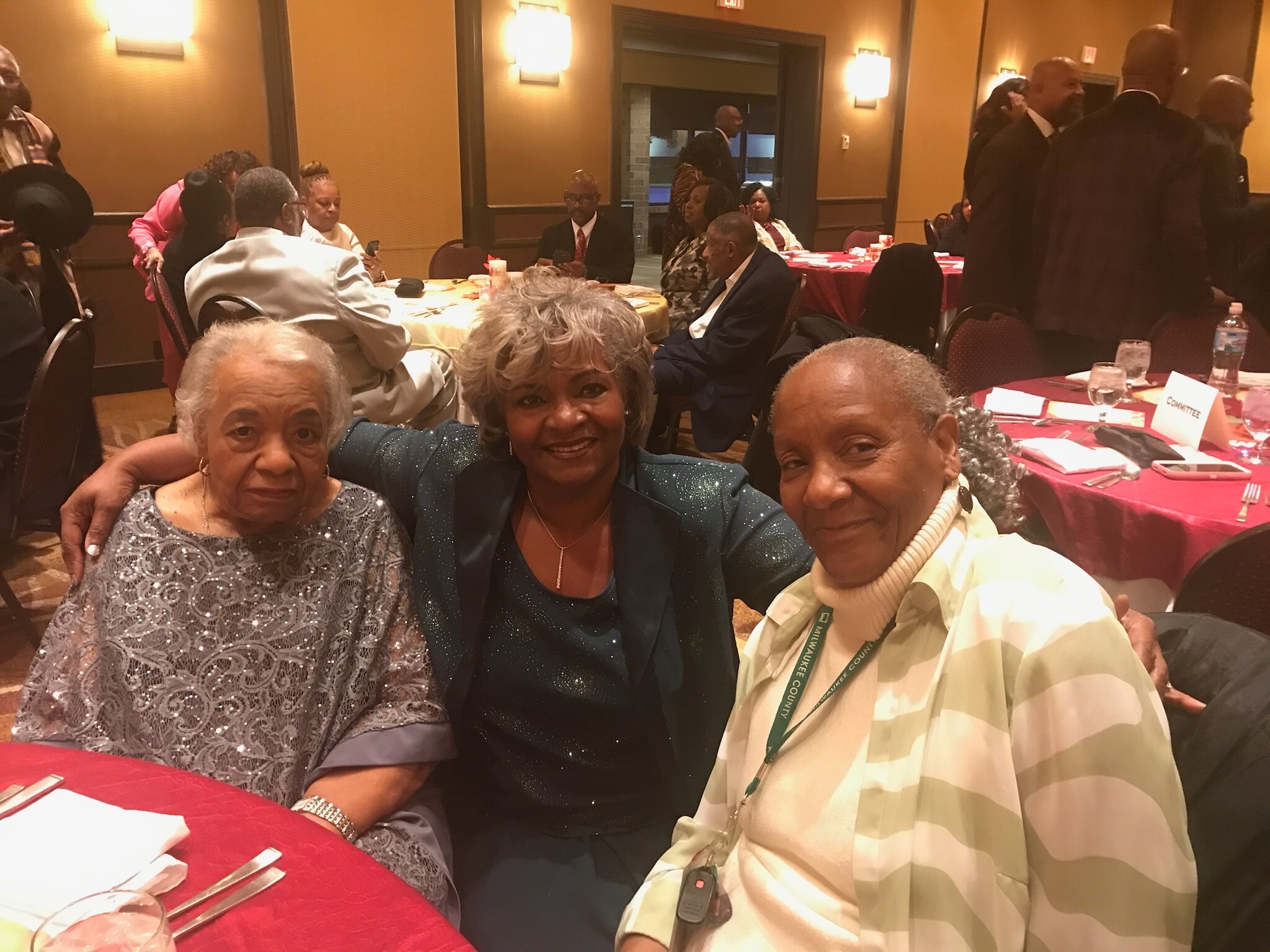 Three smiling women seated at a round banquet table in a hotel ballroom, posing for a photo in formal attire.