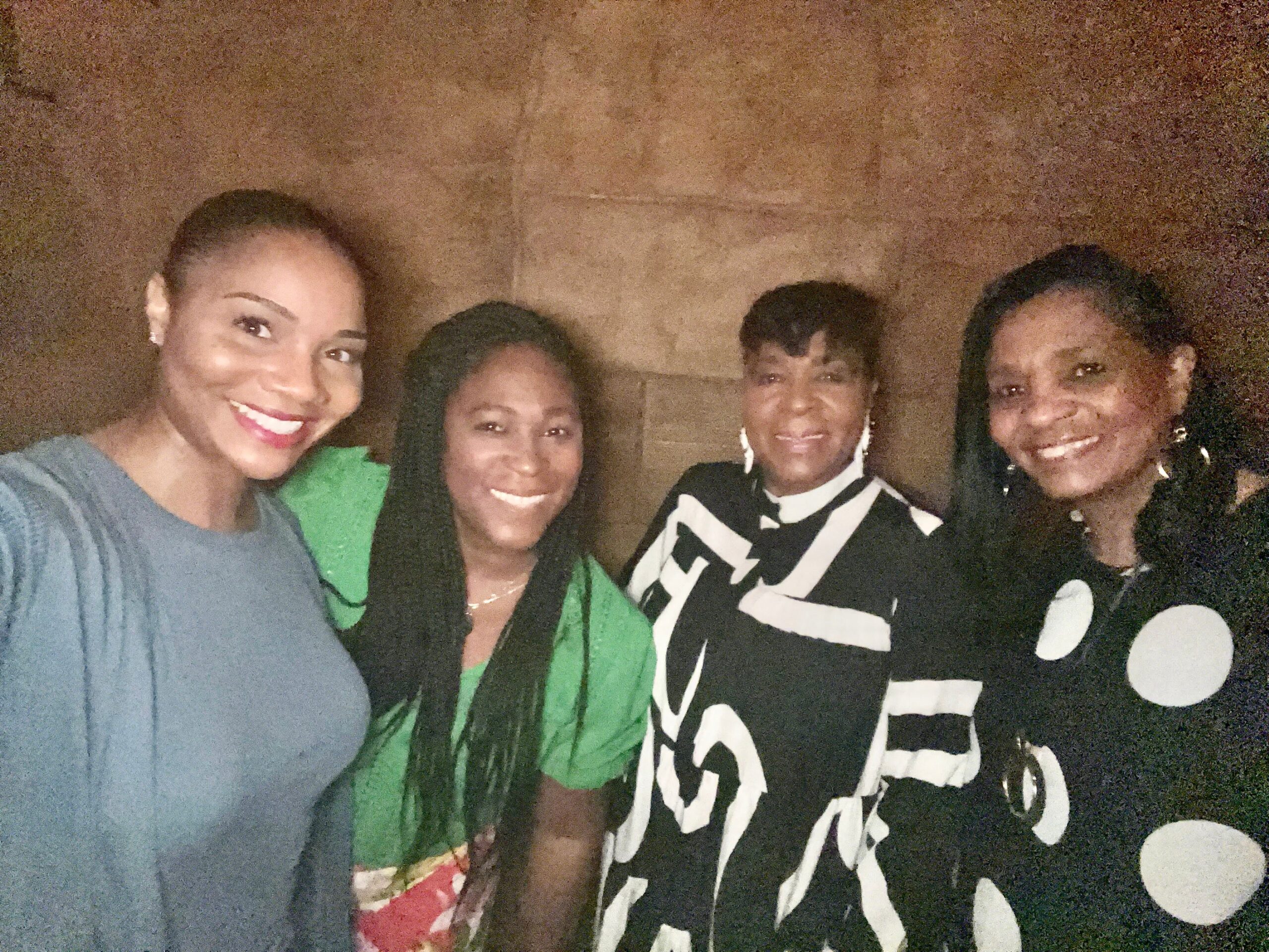 Four women taking a group selfie indoors, smiling at the camera against a wood-paneled wall.