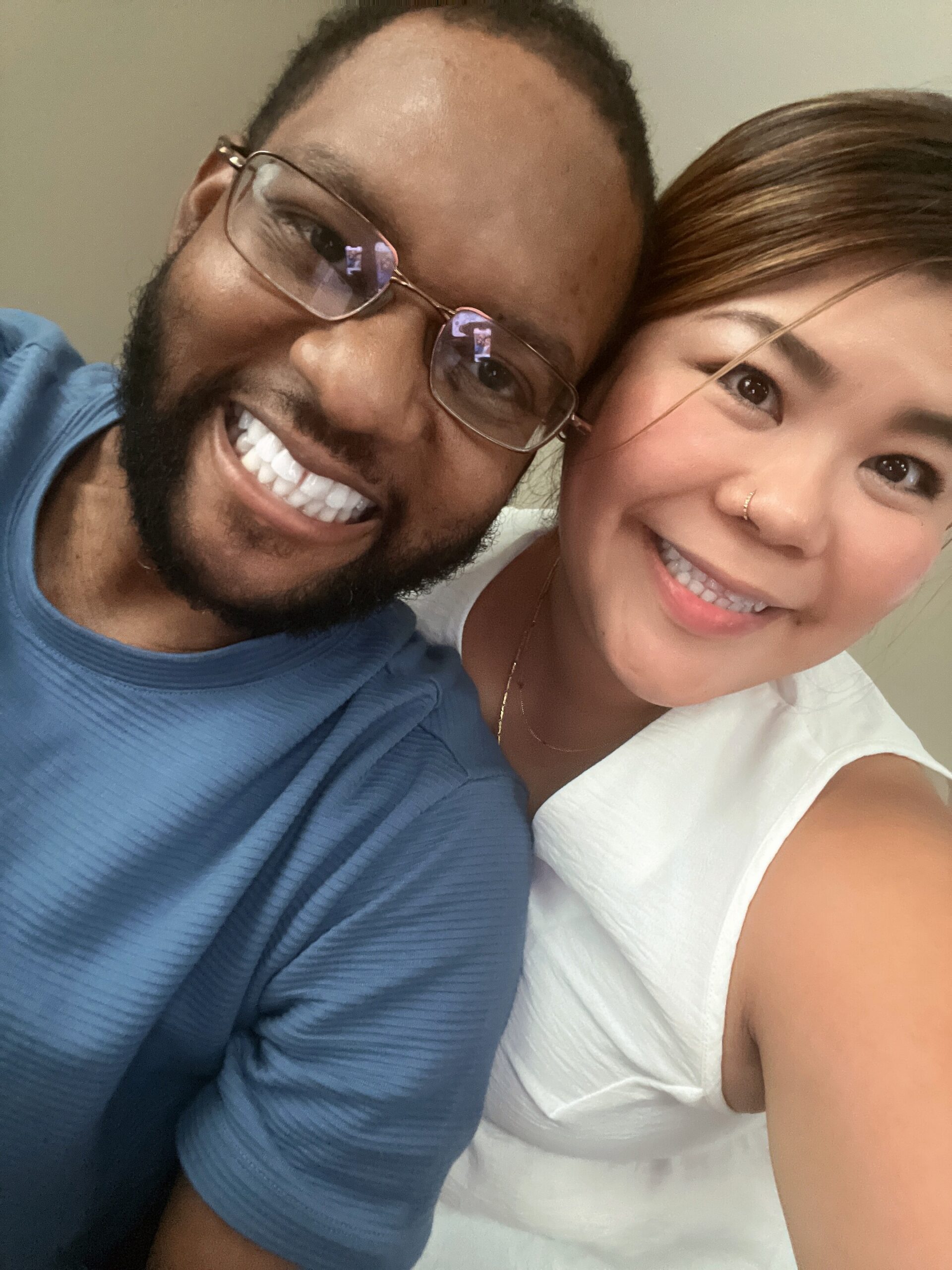 Close-up selfie of a smiling couple leaning their heads together; man wears glasses and a blue shirt, woman wears a white top and has a nose ring.