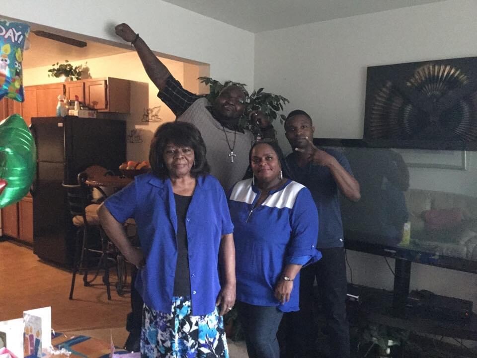 Group of four adults posing for a casual indoor party photo, two women in blue shirts in front, a man raising an arm behind them, balloons and a kitchen in the background.