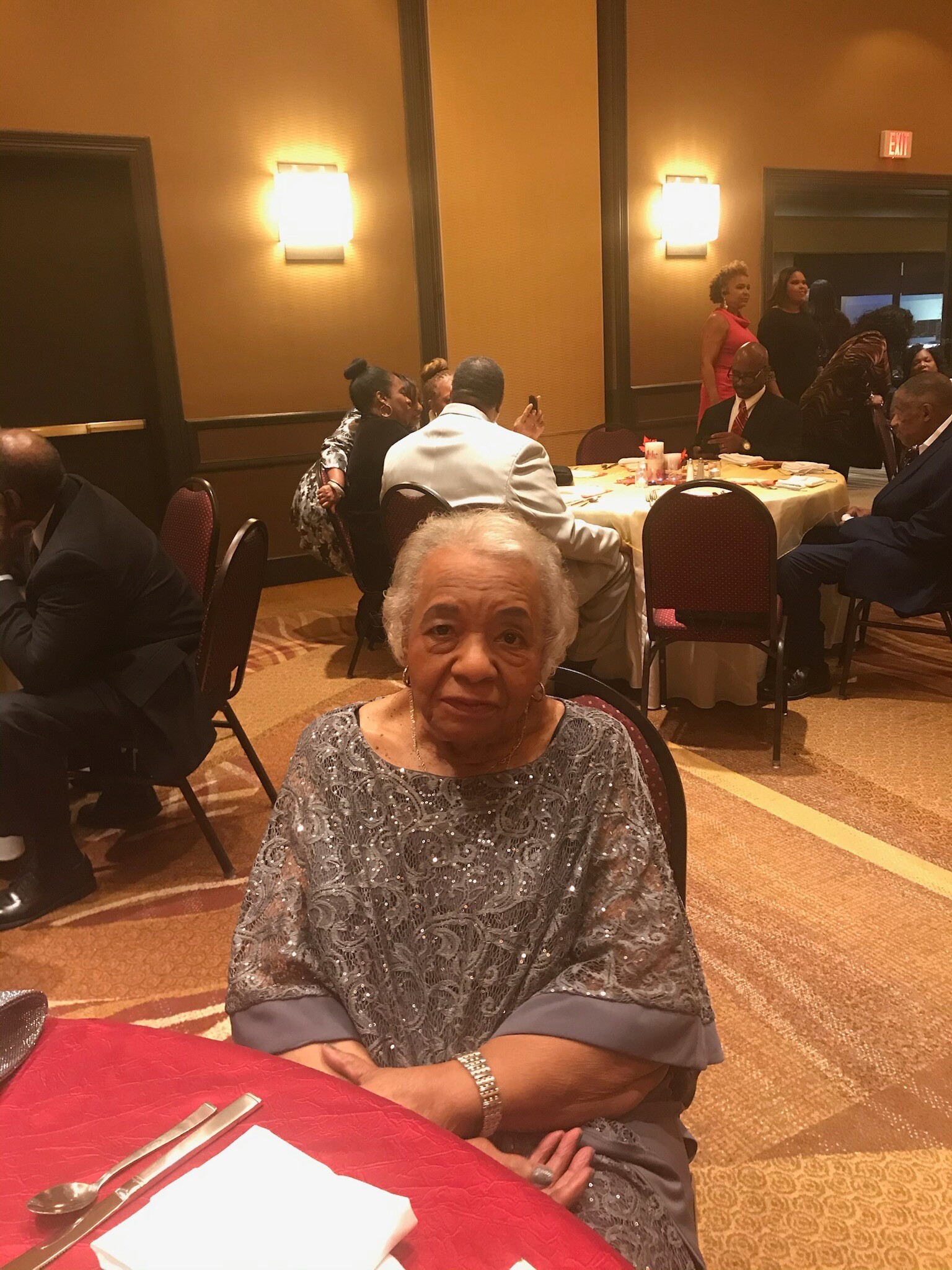 Elderly woman in a silver lace dress seated at a banquet table in a formal event hall.