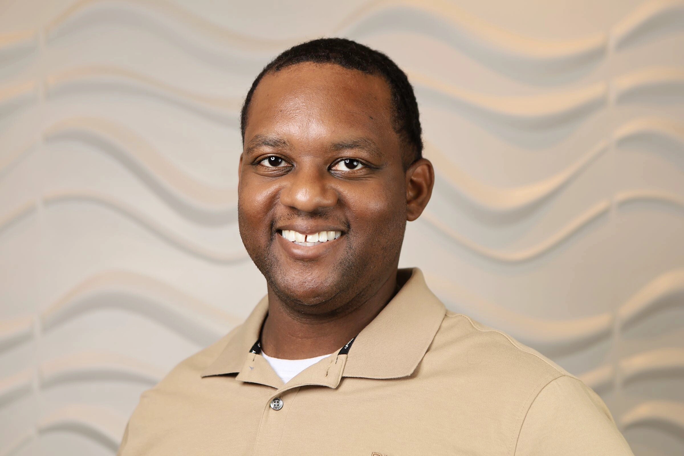 Smiling man wearing a tan polo shirt against a light wavy-patterned background.