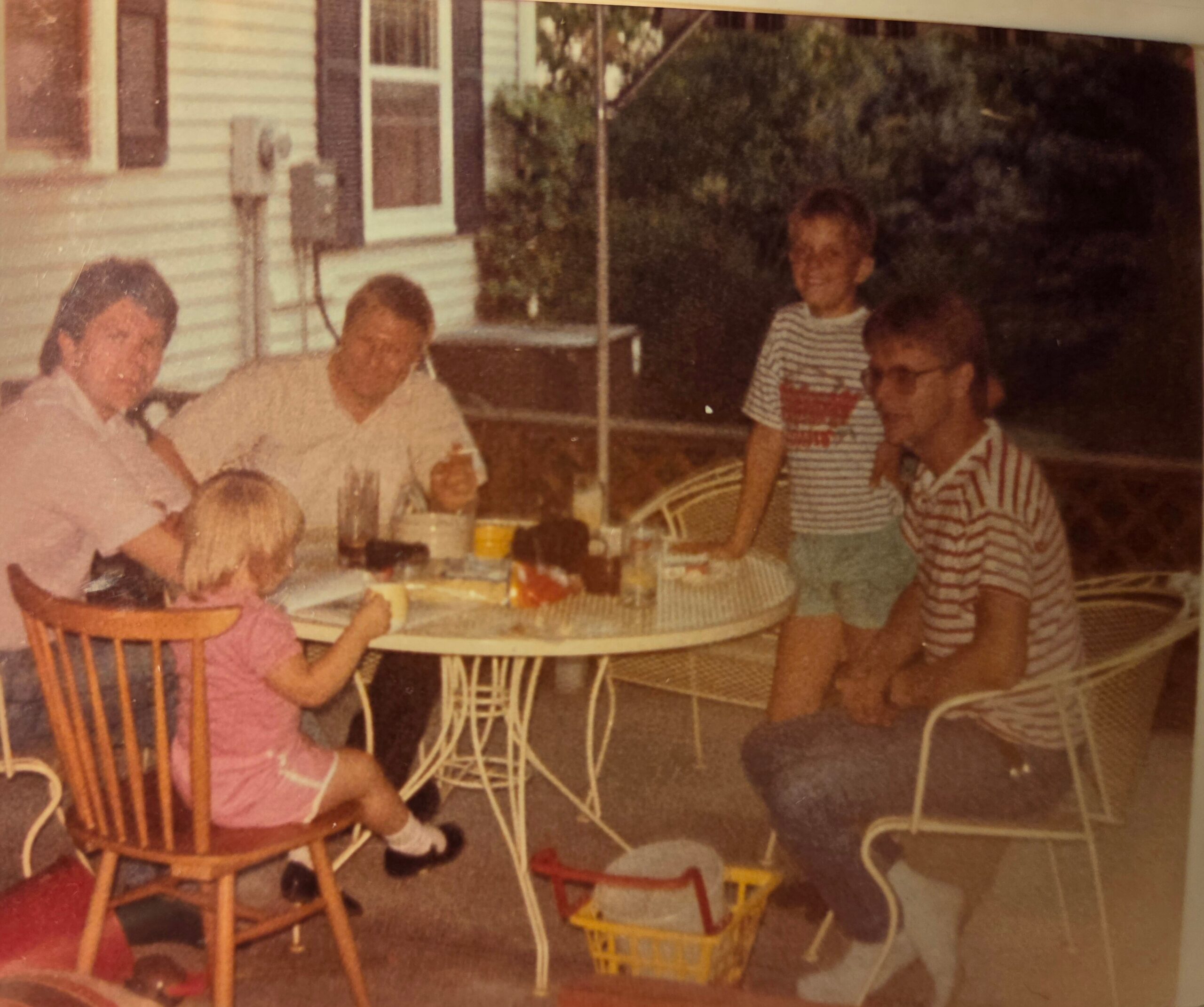 Group of children and adults at a round table on a backyard patio, with cups and snacks visible.