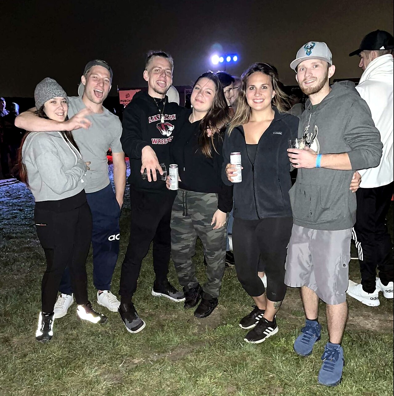 Group of six friends posing outdoors at night, smiling, with drinks in hand and arms around each other on a grassy field.