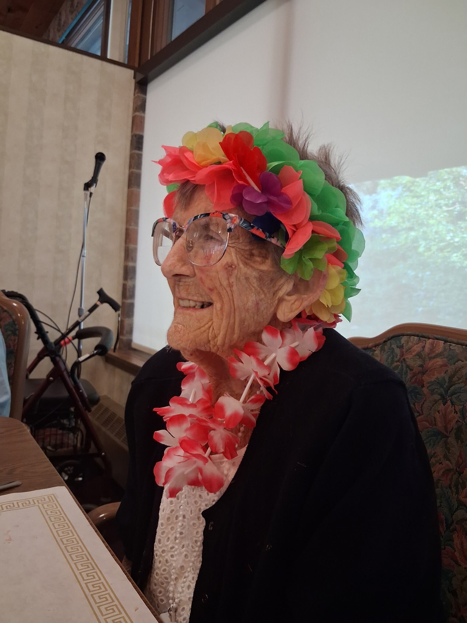 Older woman wearing a bright multicolor flower lei and headband, with glasses, seated at a table during a celebration.