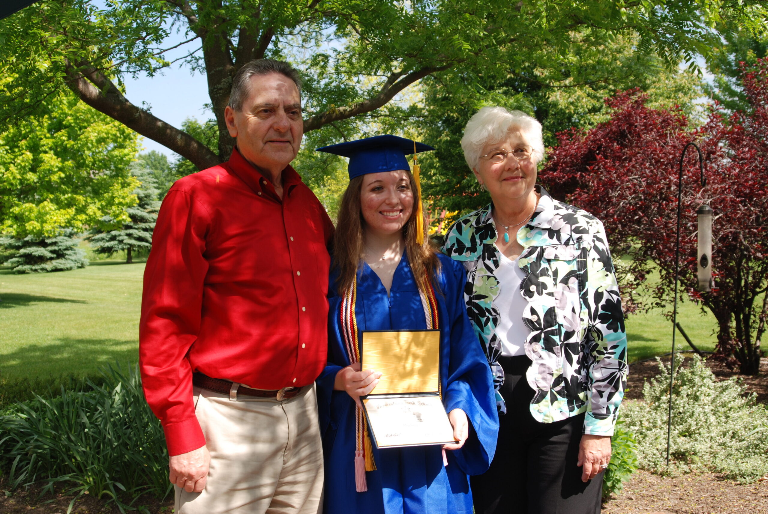 Graduate in blue cap and gown with two smiling adults outdoors in a sunny park, holding a diploma folder.