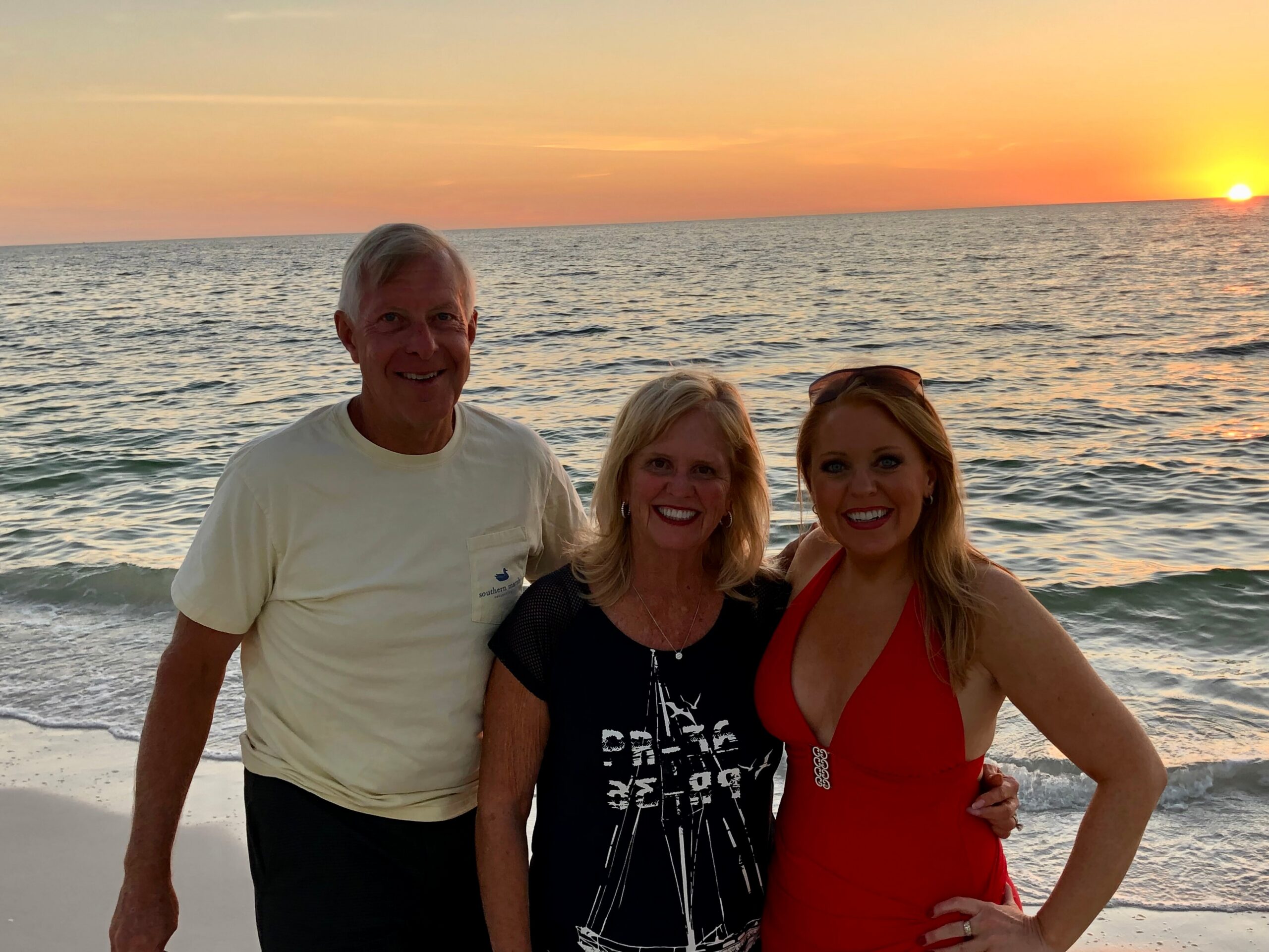 Three people posing on a beach at sunset, with orange sky and calm waves in the background (man in a pale shirt on the left, two women in the center and right).