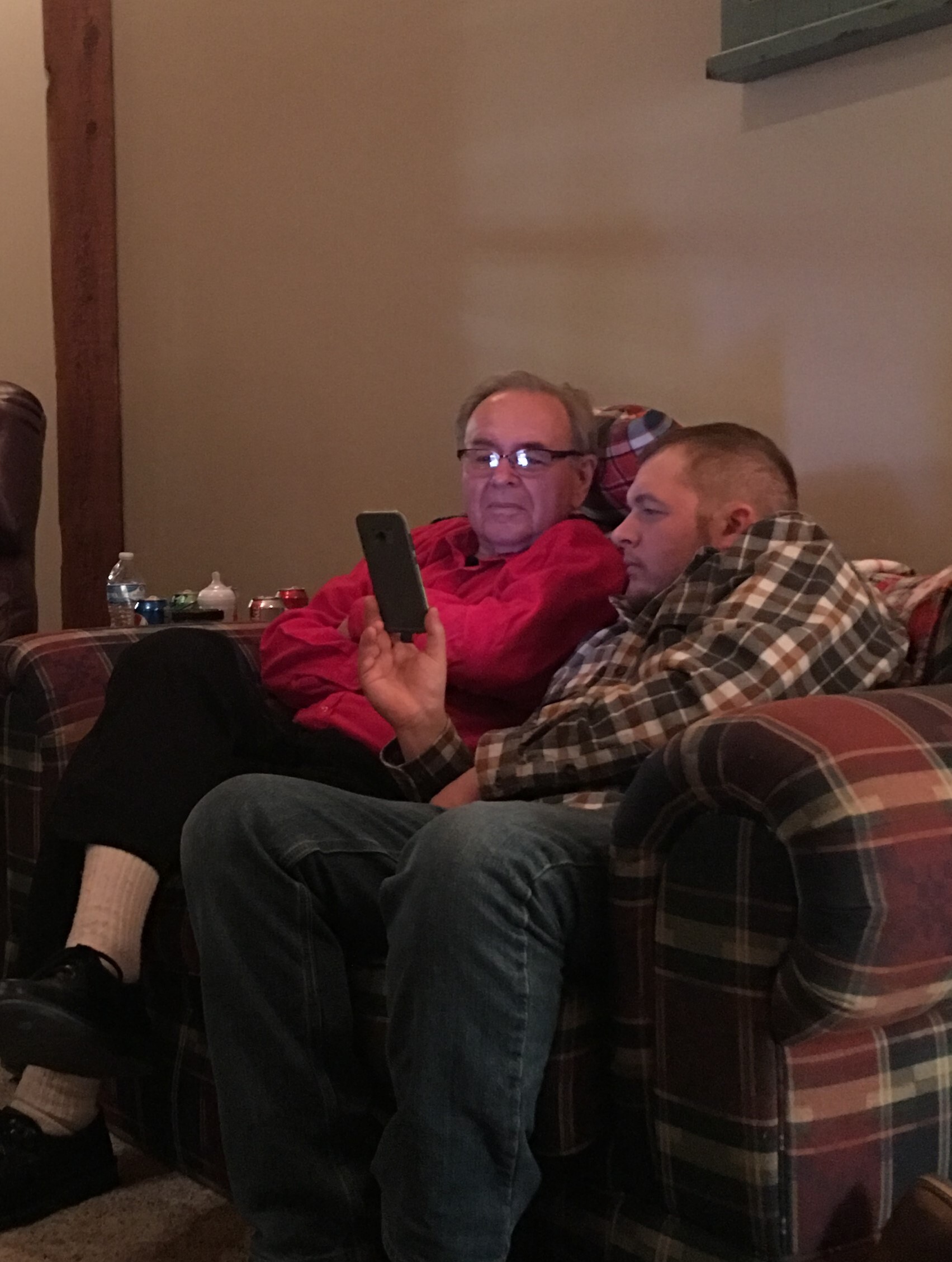 Two men on a plaid sofa indoors; one in a red jacket using a smartphone, the other wrapped in a plaid blanket.