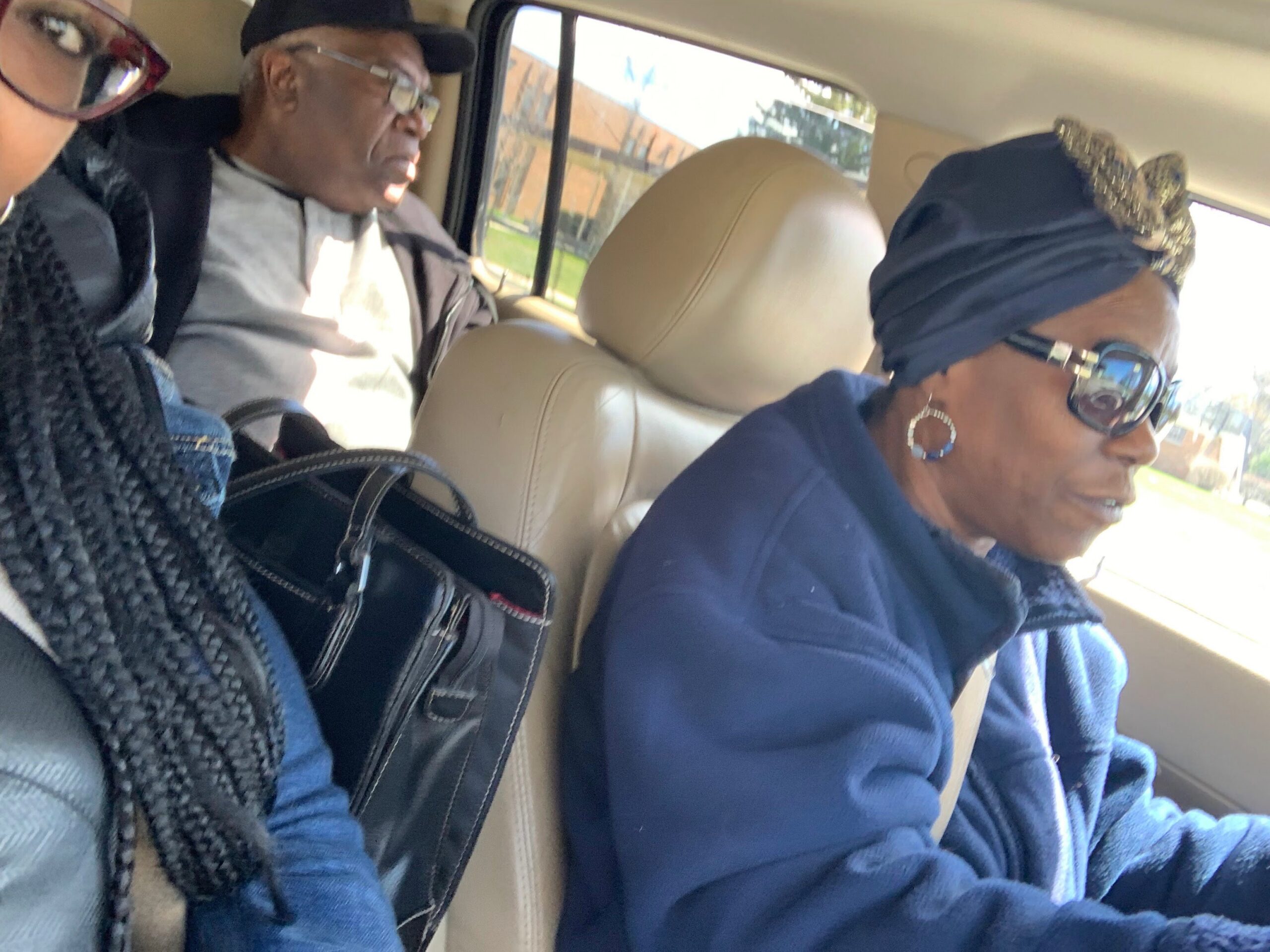 Three adults riding in a beige car: a woman in the front passenger seat wearing a navy head wrap and sunglasses, a man in the back seat with a cap and glasses, and a person with braided hair partially visible on the left.
